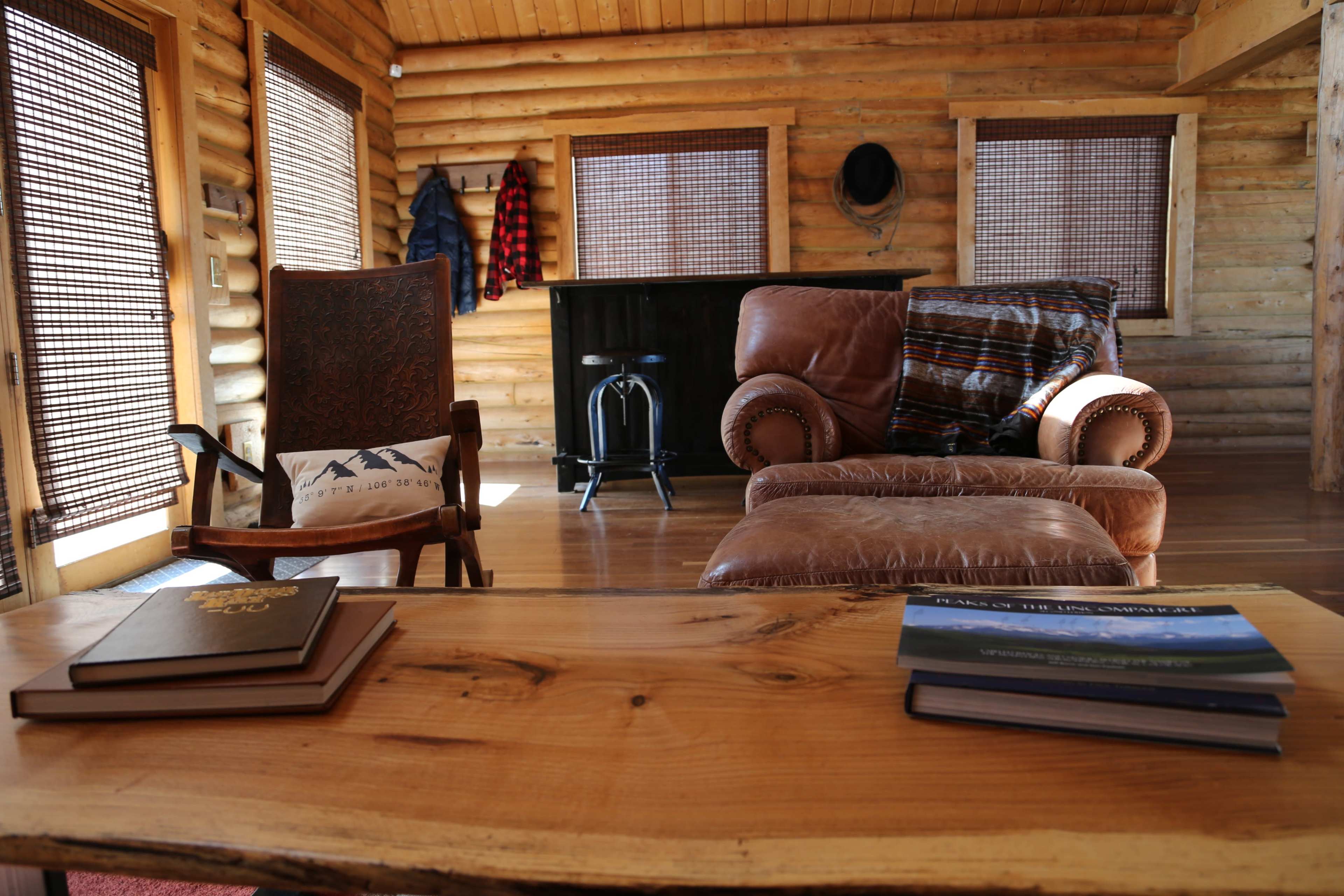 The room features a wooden table with books, two chairs, and a bar area, all set within a log cabin interior.