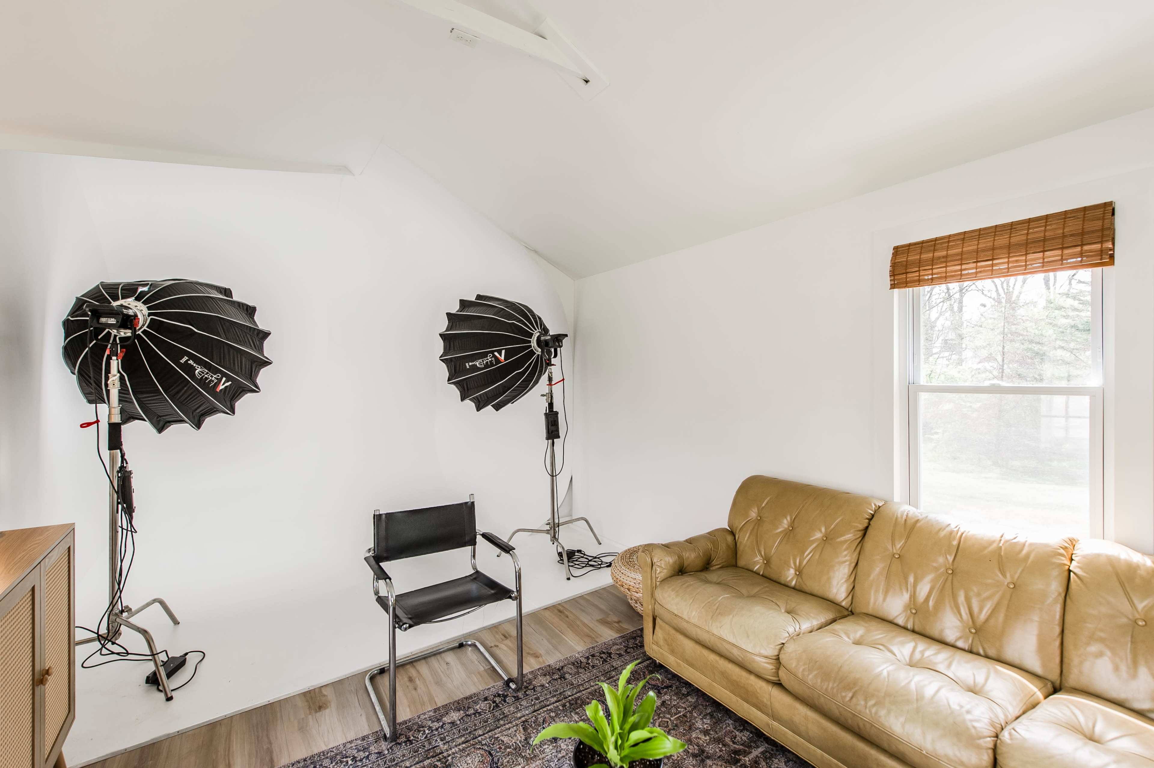 A well-lit room features two photography light setups and a tan leather sofa near a window.