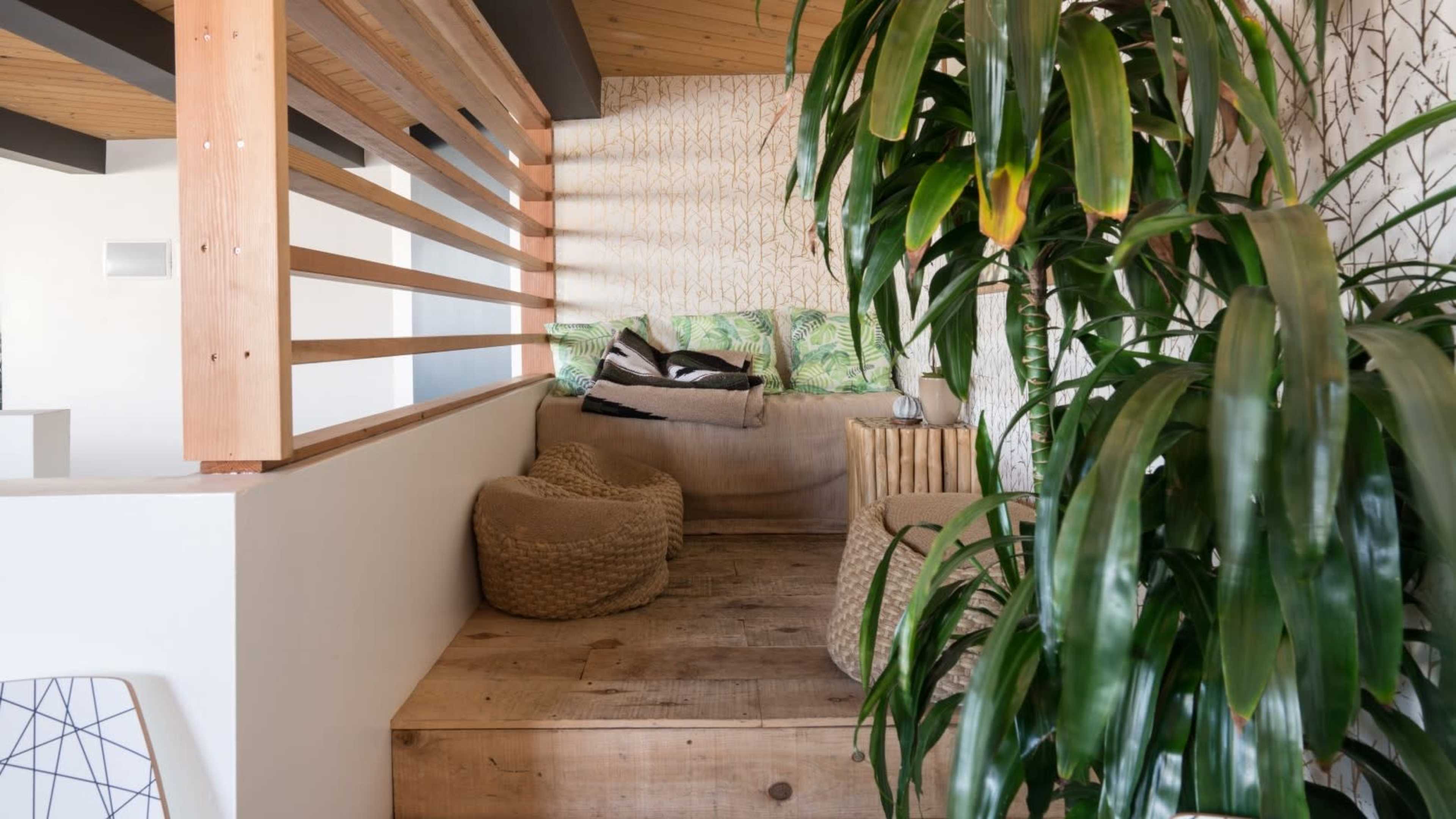 The image shows a cozy nook with a wooden platform, surrounded by plants, featuring cushions and a side table.