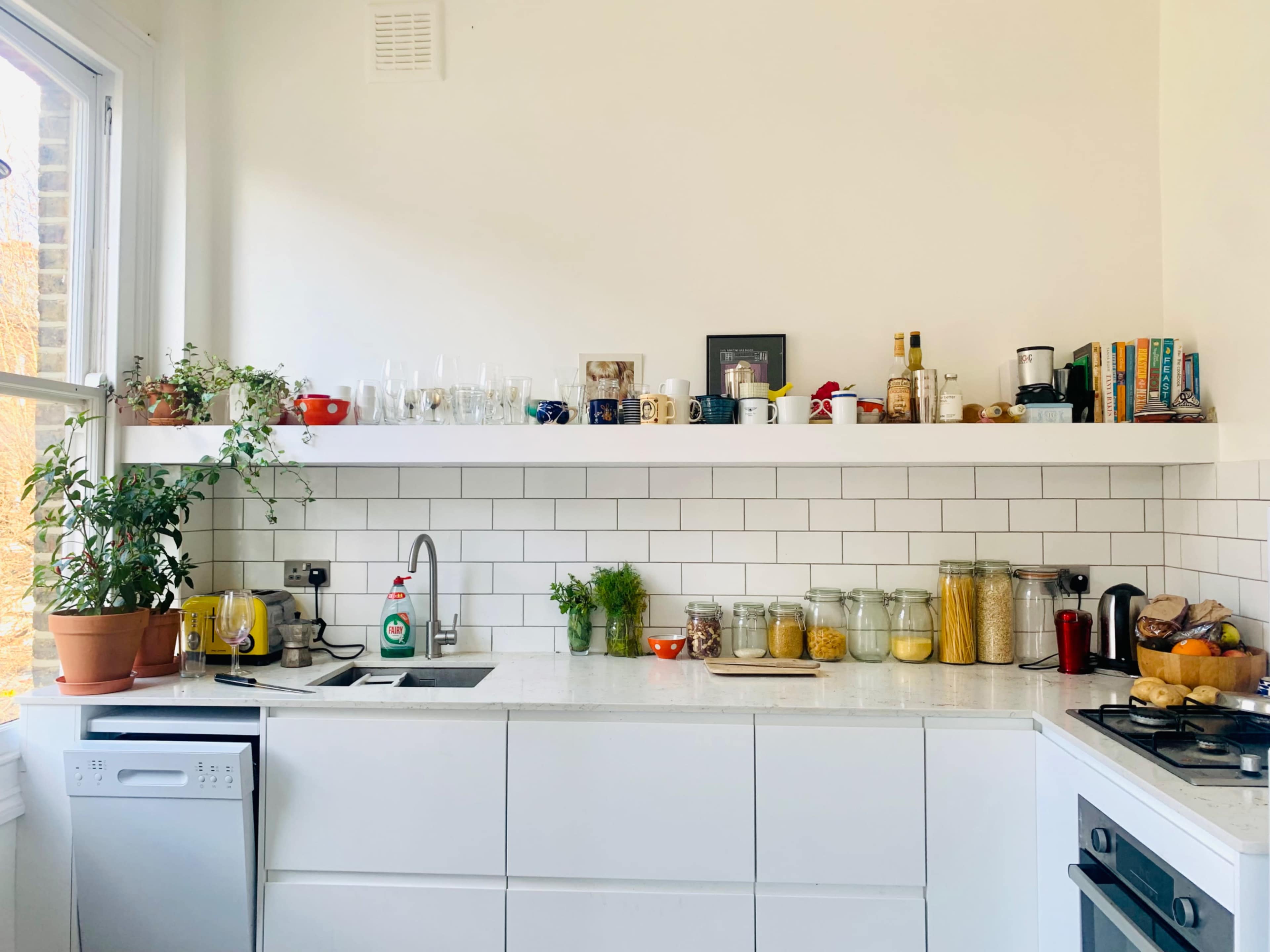 A modern kitchen features a white countertop with a sink, a dishwasher, various jars of food, and a shelf displaying glassware and kitchenware.