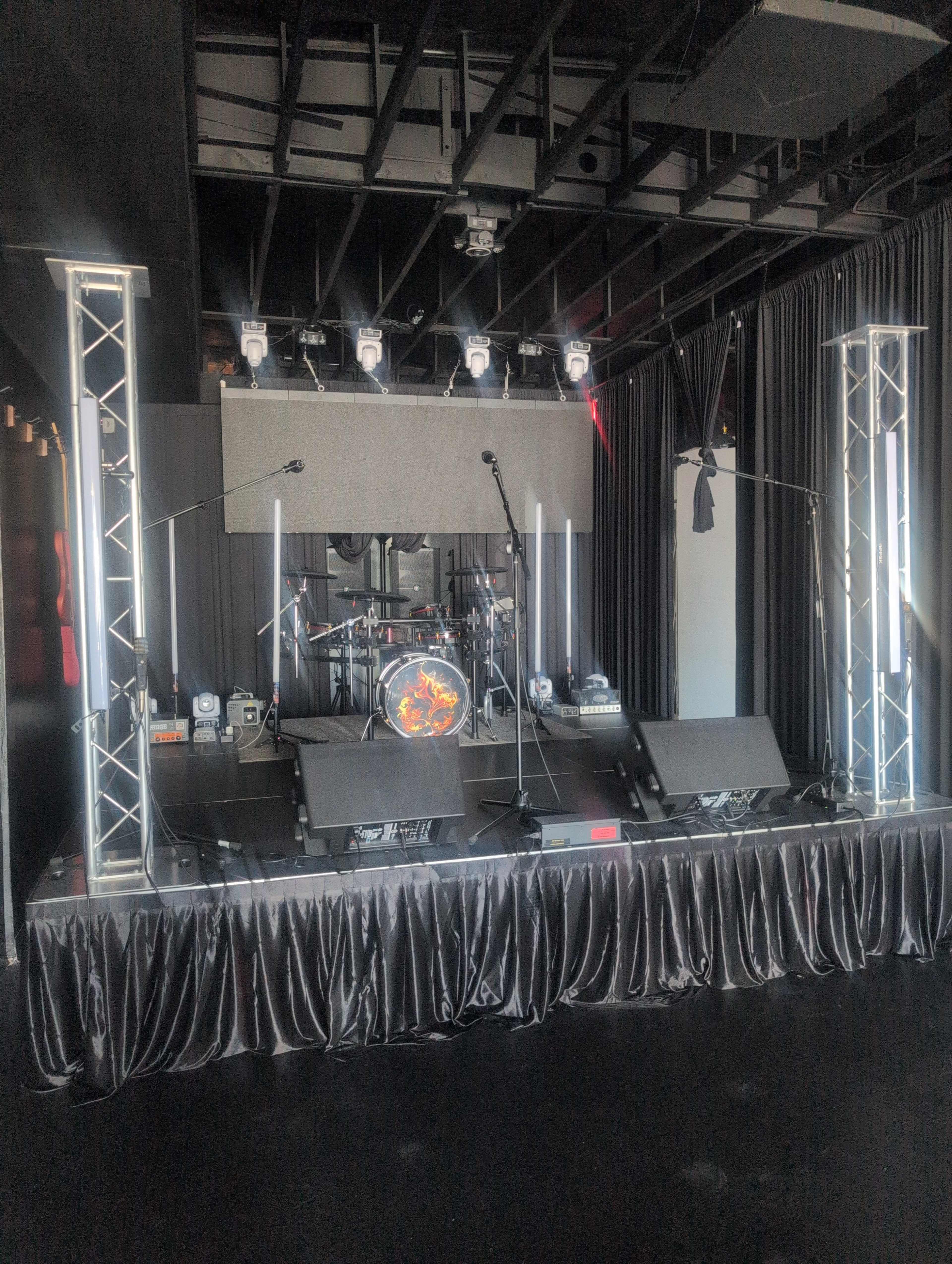 The image shows a stage setup with a drum kit, microphones, and lighting fixtures in a dimly lit venue.