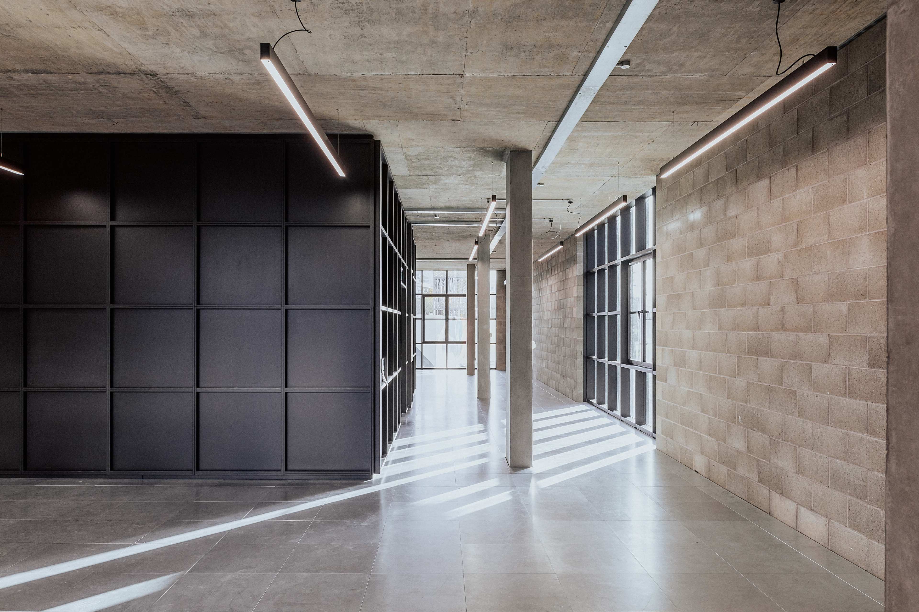 The image shows a modern interior space with a concrete ceiling, large windows, and walls featuring a combination of black paneling and light-colored masonry.
