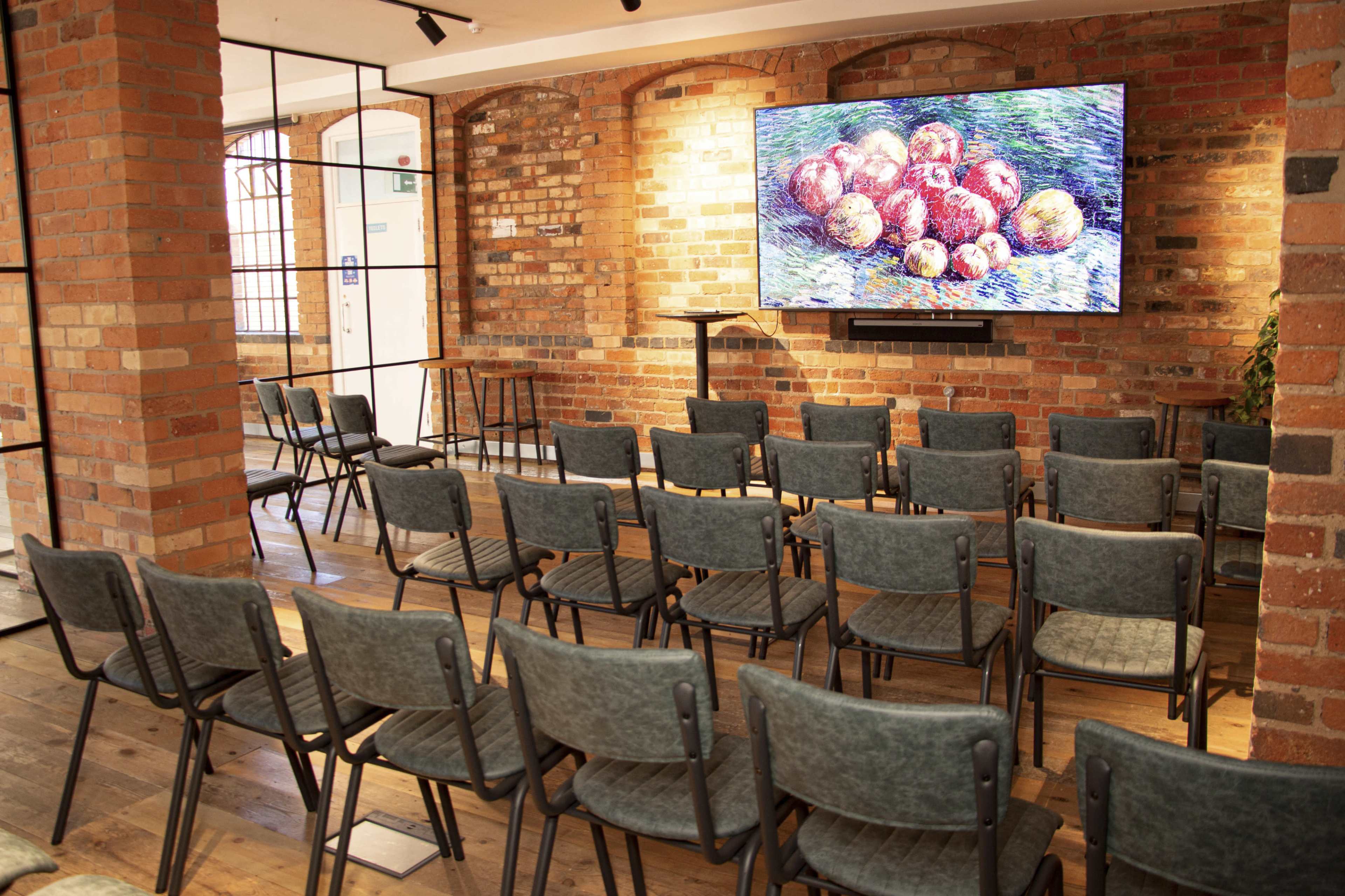 A room with empty chairs arranged in rows facing a large television screen displaying a still life painting of apples, with brick walls and wooden flooring.