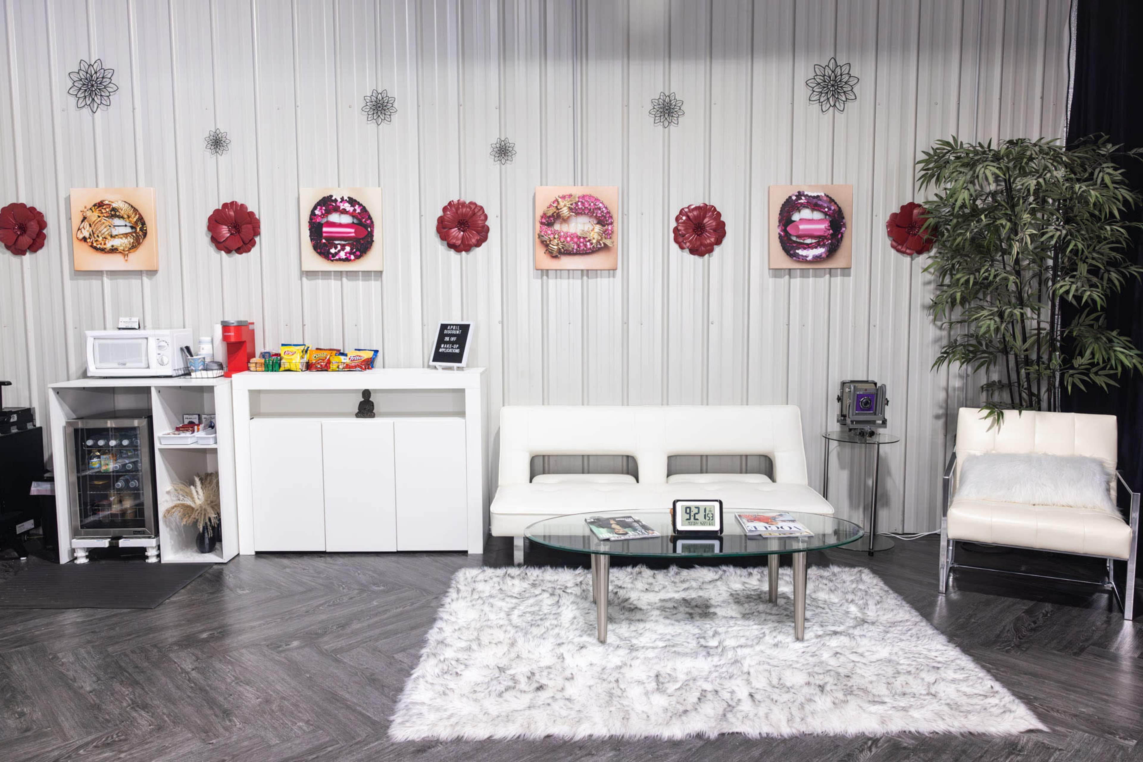 A modern lounge area with a white sofa, glass coffee table, and a small kitchenette, decorated with colorful wall art and floral accents.