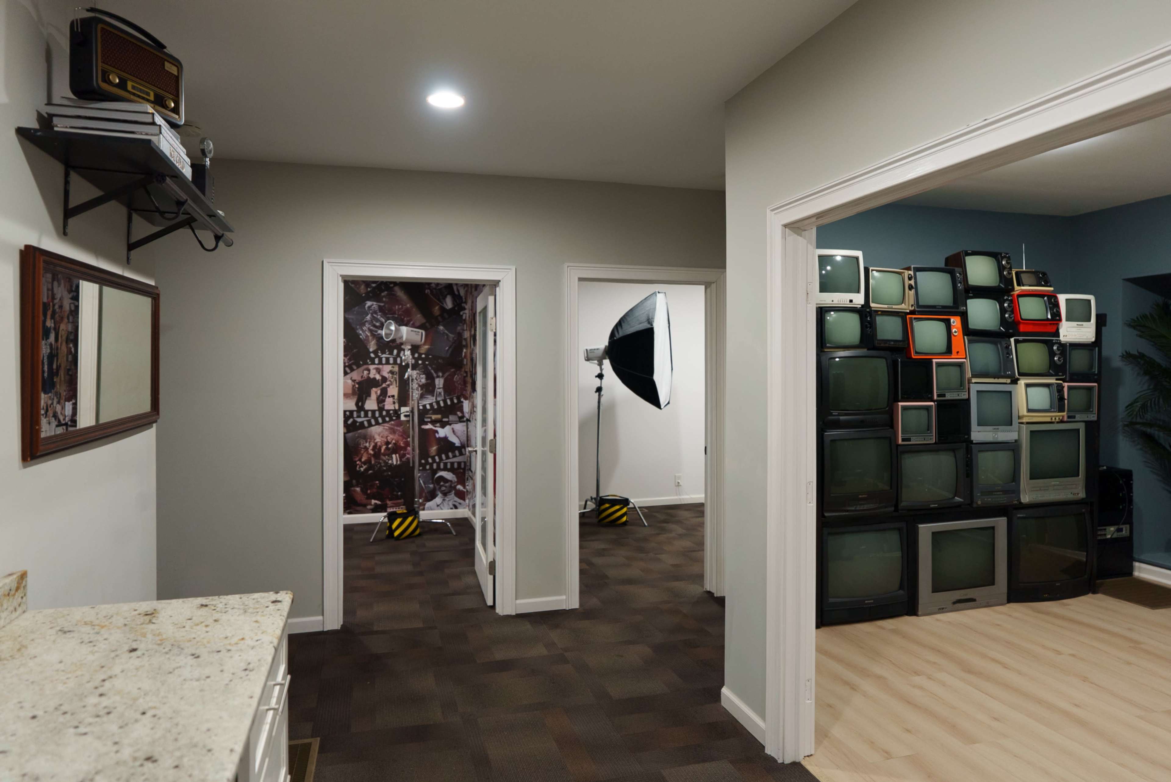 The image shows a room with a countertop on the left, a mirror on the wall, and a hallway leading to a room filled with stacked vintage televisions and a photography light setup.