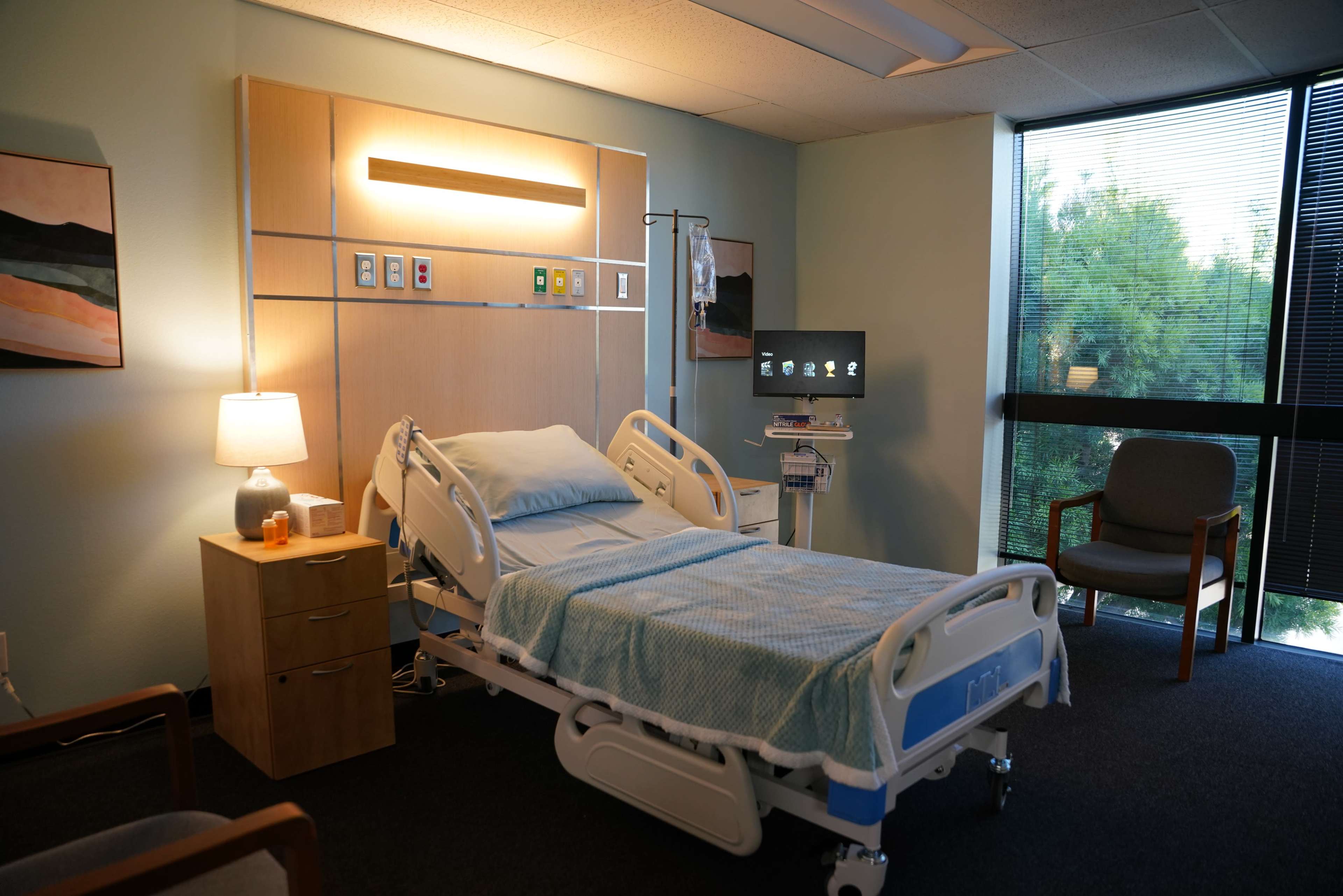 The image shows a hospital room featuring a bed, a nightstand, a chair, and medical equipment, with large windows allowing natural light to enter.