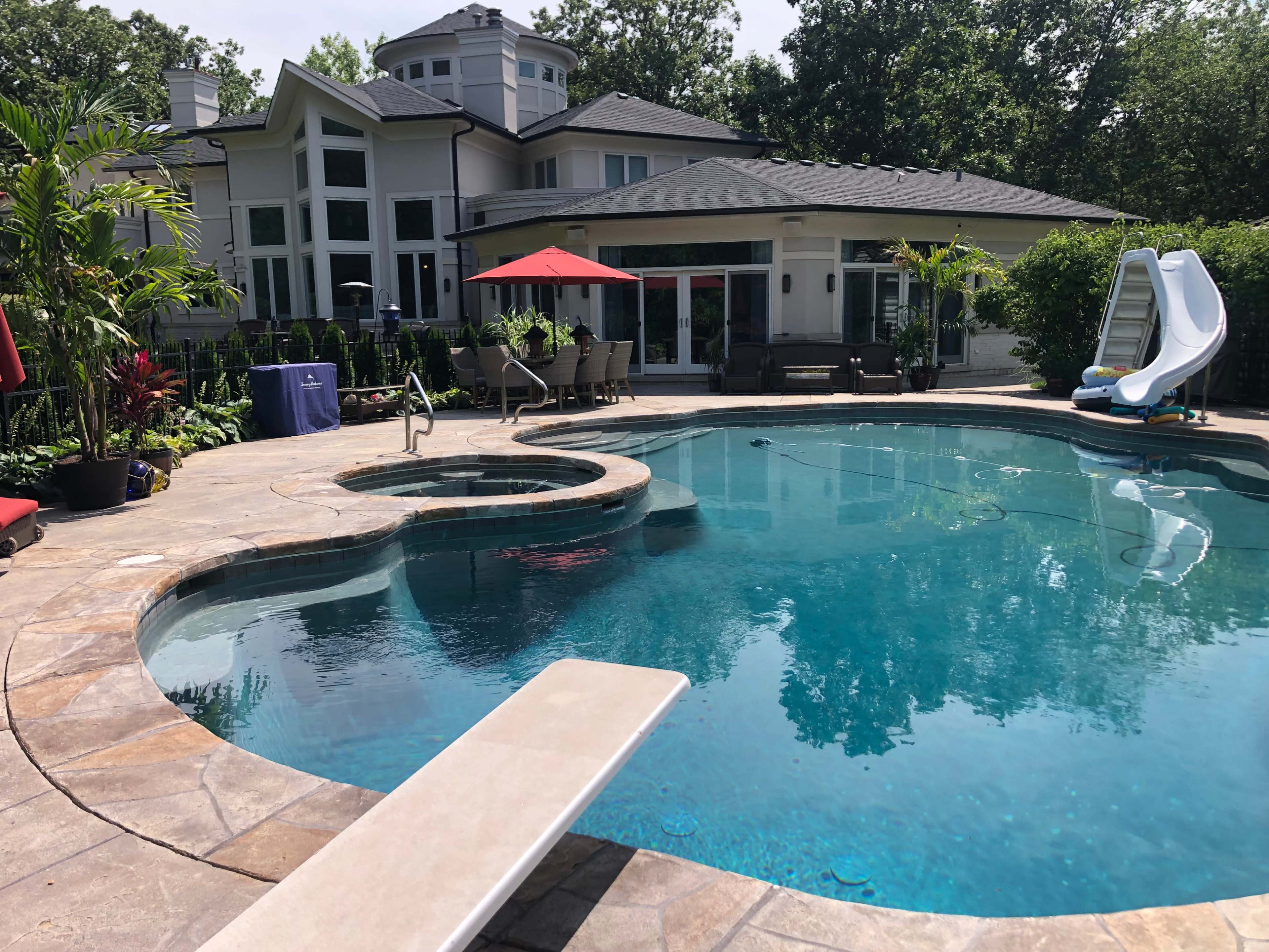 A residential backyard with a swimming pool, a diving board, and a waterslide beside a large house surrounded by greenery.