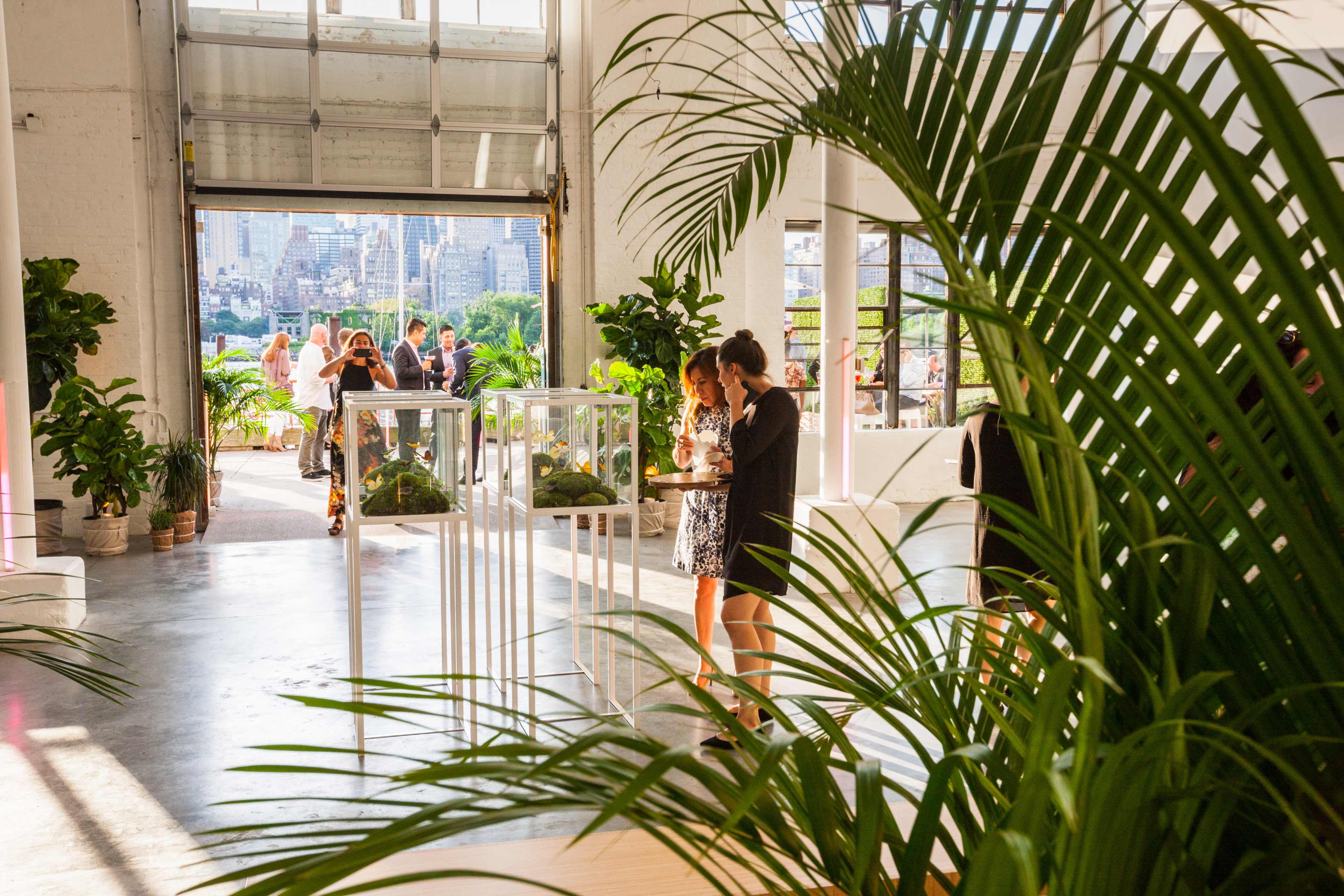 The image shows an indoor event space with large windows, featuring guests mingling around display cases, surrounded by greenery.