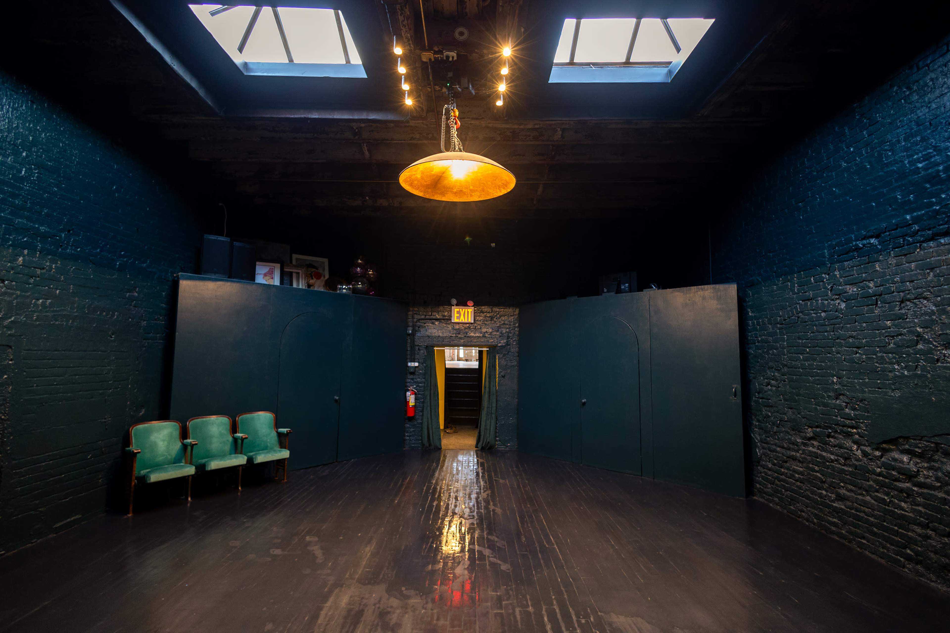 A spacious, dimly lit room with dark walls, wooden floors, and a large fixture hanging from the ceiling, featuring two green chairs positioned against one wall.