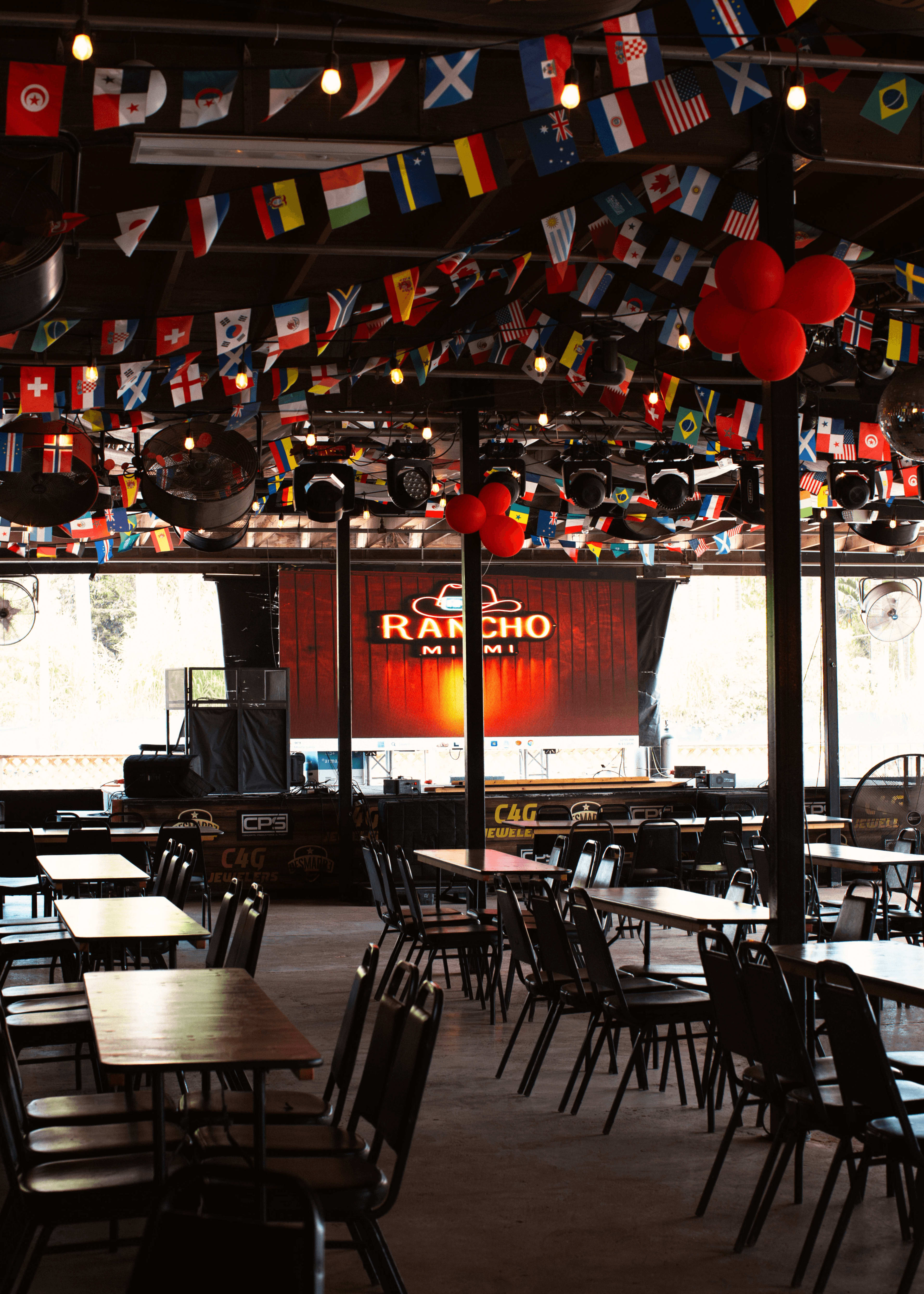 The interior of a venue features tables and chairs arranged beneath a ceiling adorned with hanging flags from various countries, with a stage lit in the background.