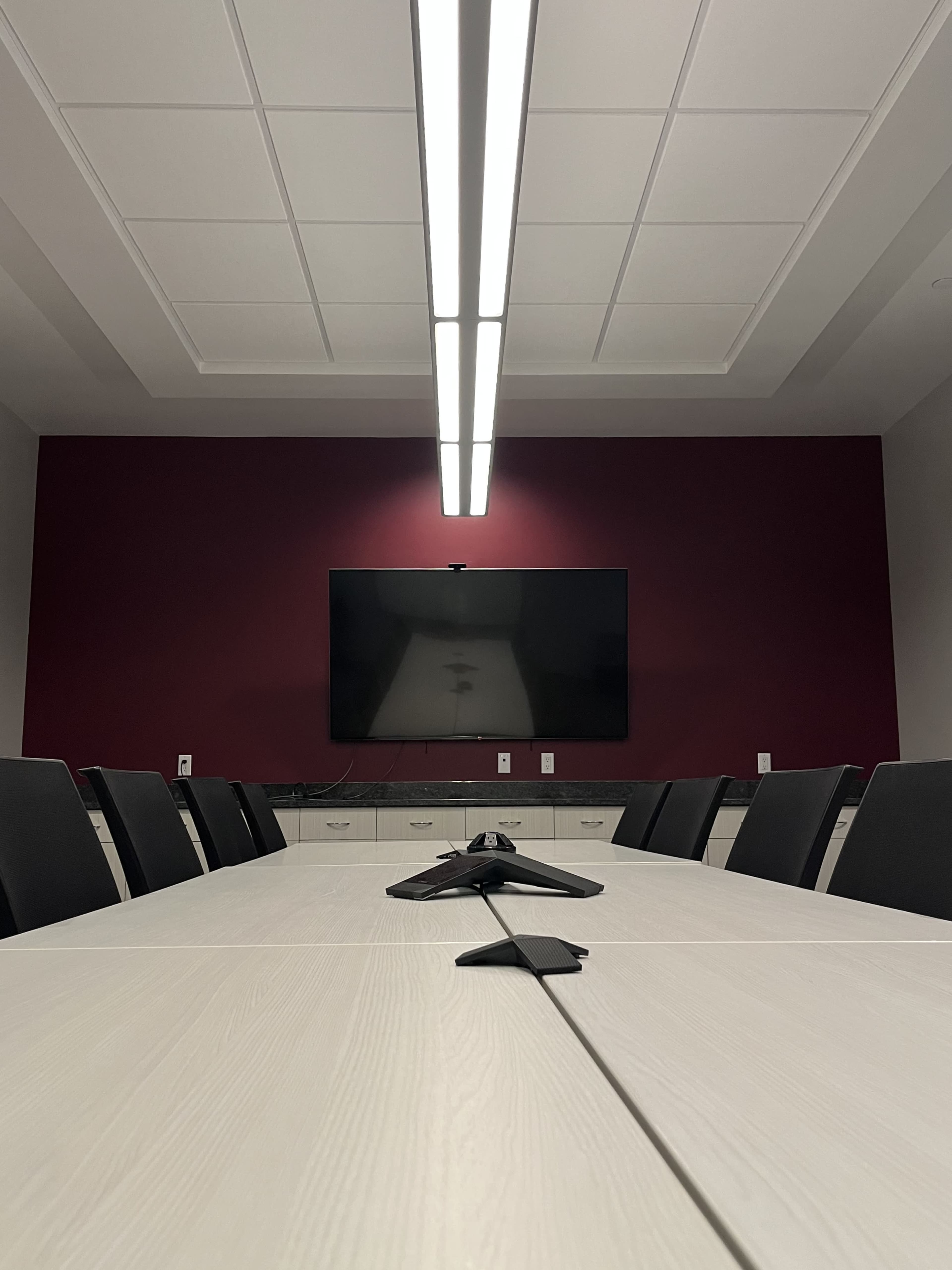 A modern conference room features a long table, black chairs, and a flat-screen TV mounted on a burgundy wall.