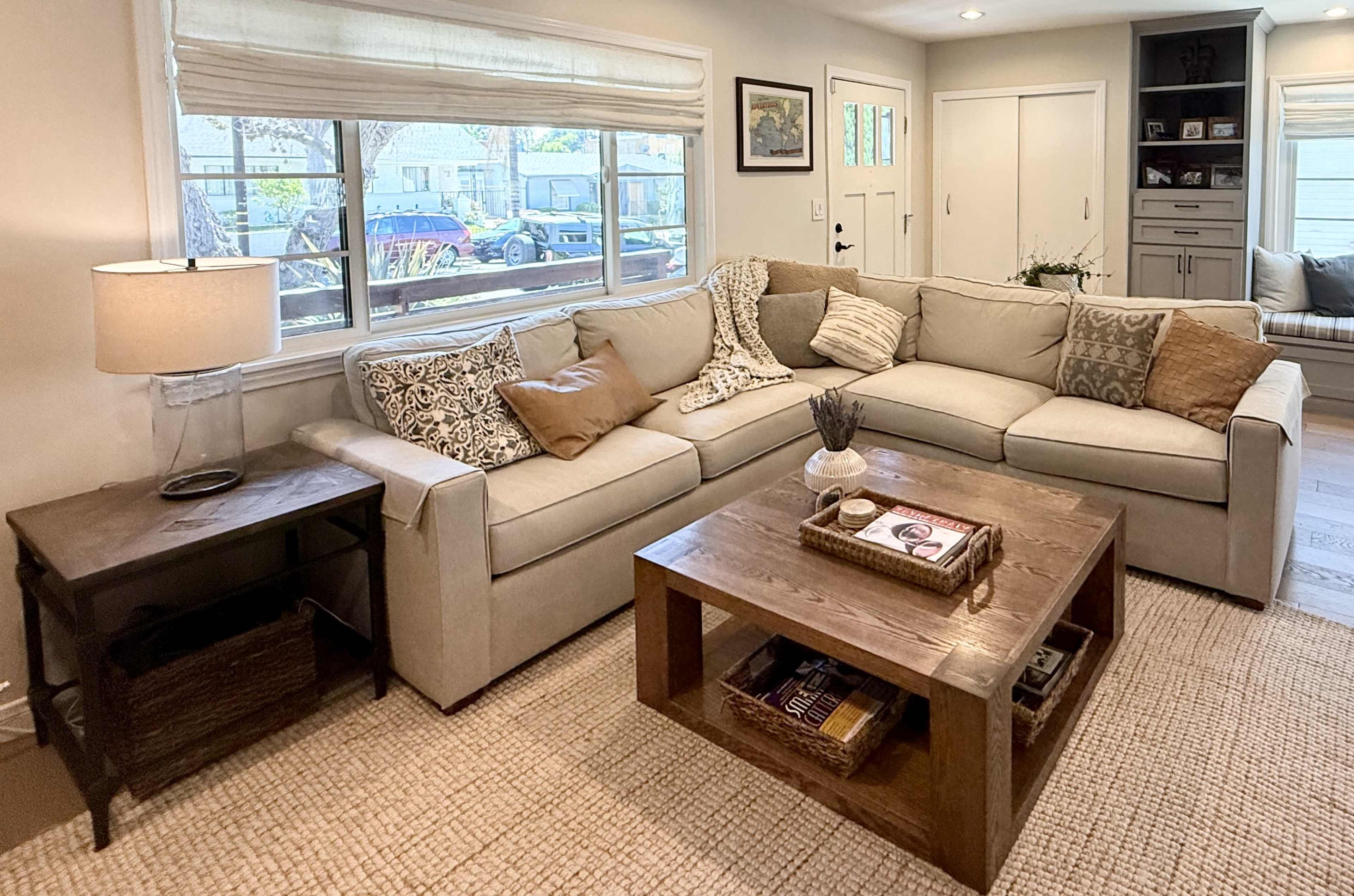 A cozy living room features a light-colored sectional couch adorned with various decorative pillows, a wooden coffee table, and a side table with a lamp, all arranged near a window.