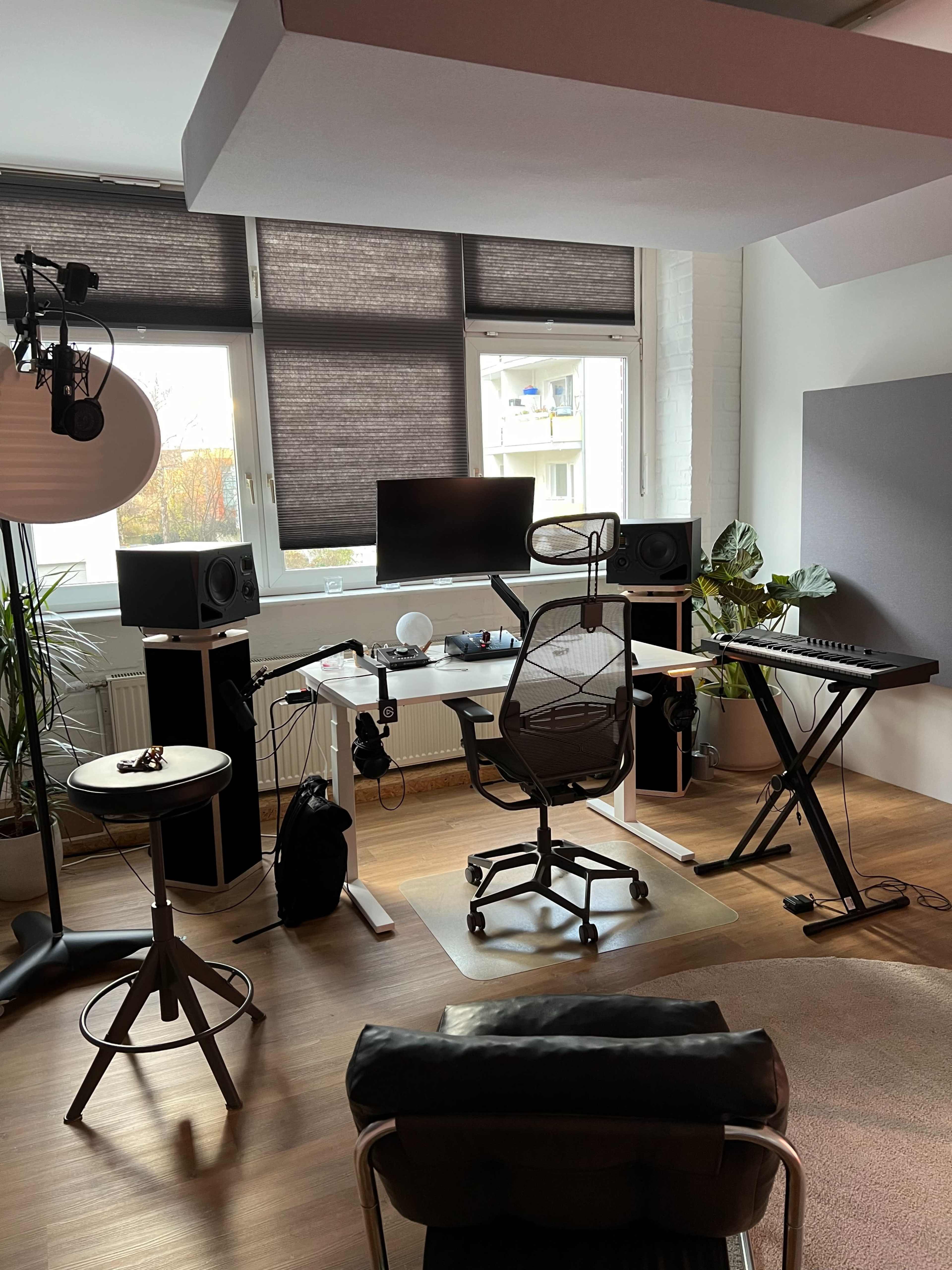 The image shows a music production studio featuring a chair, desk with a computer monitor, audio speakers, and a keyboard, accompanied by a plant and large windows.
