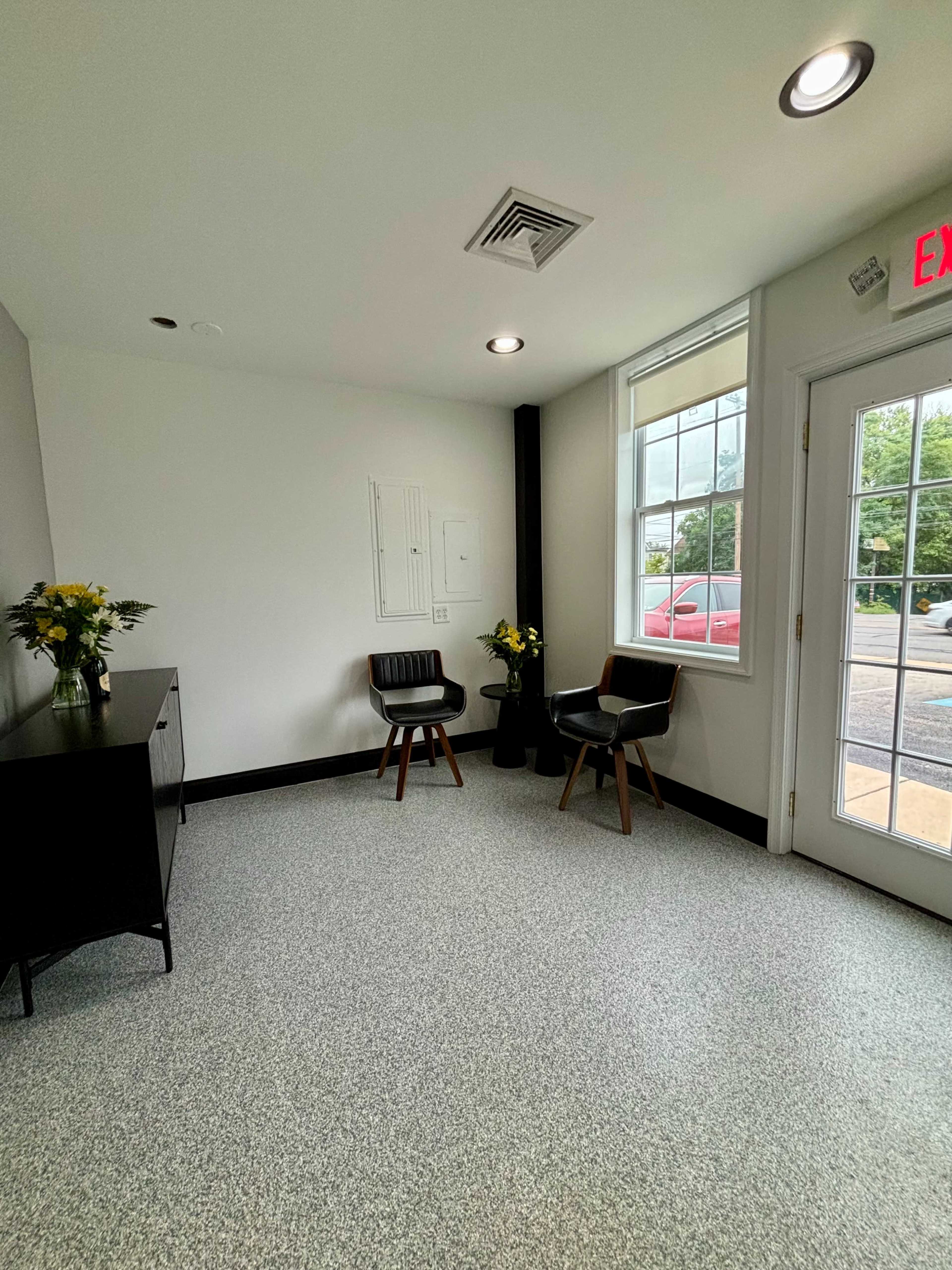 The image shows a small waiting area with two black chairs, a side table with a vase of yellow flowers, and a door leading outside.
