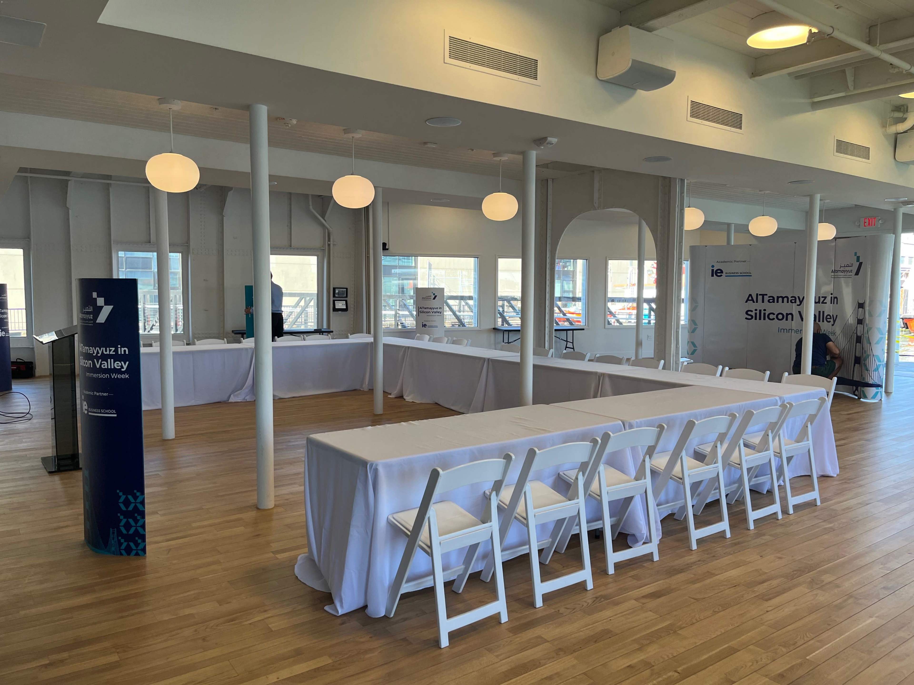 A spacious event venue with white tables and chairs set up, featuring large windows and decorative lighting fixtures.