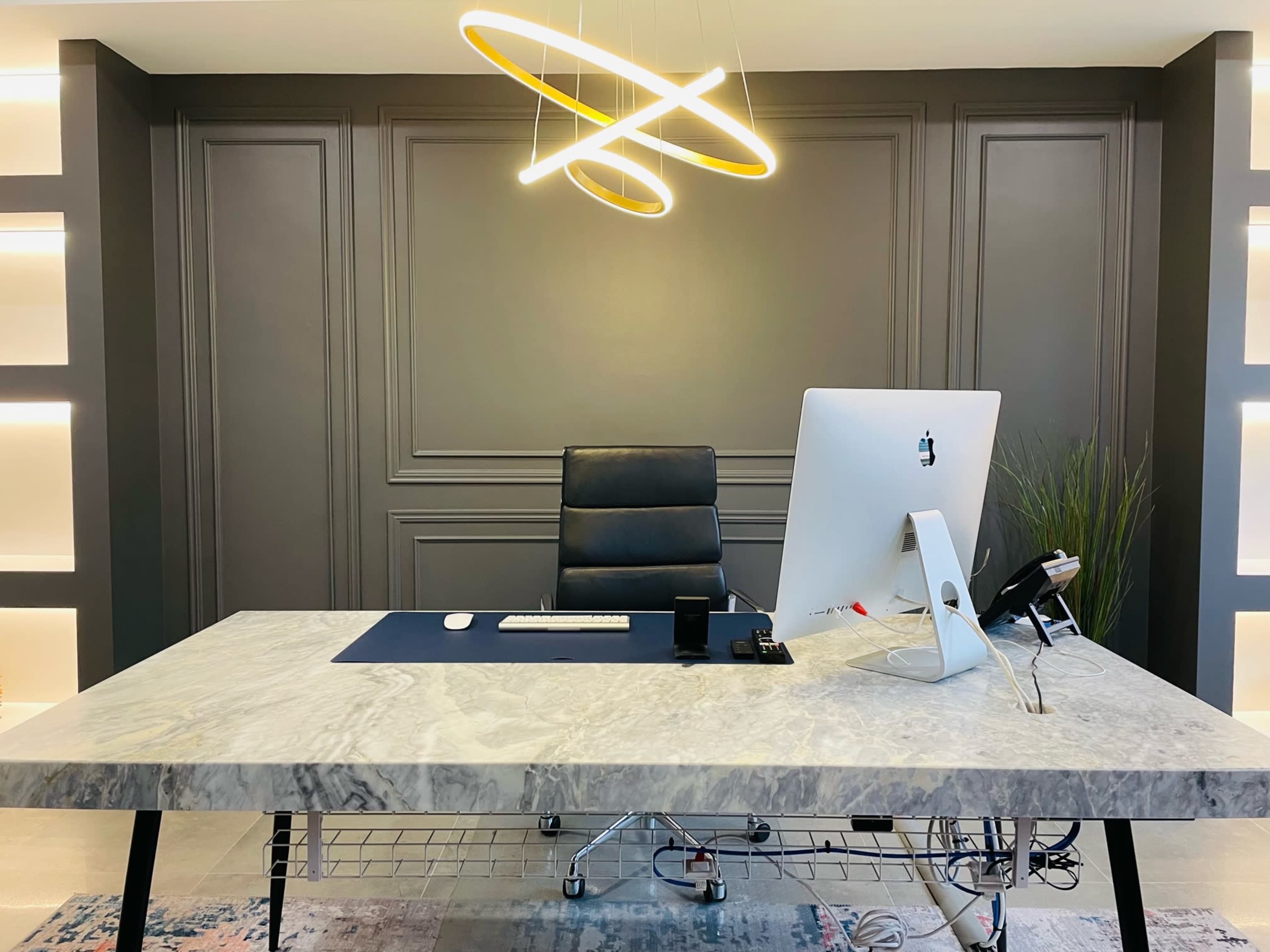 A modern office space features a sleek marble desk, a high-backed black chair, and a computer against a dark textured wall with ambient lighting.