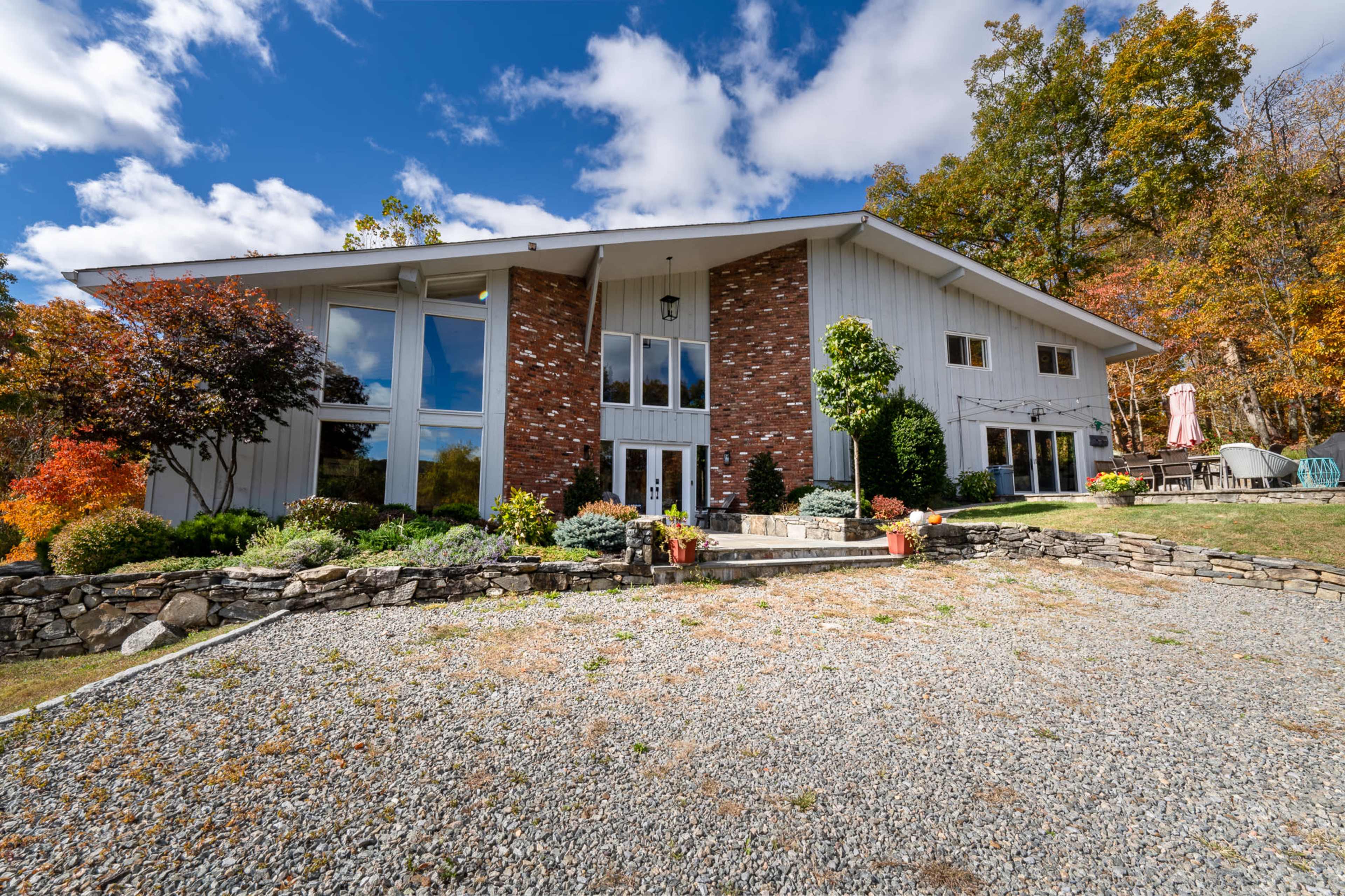 A modern two-story house with large windows and a brick accent wall is set against a backdrop of autumn foliage and a gravel driveway.