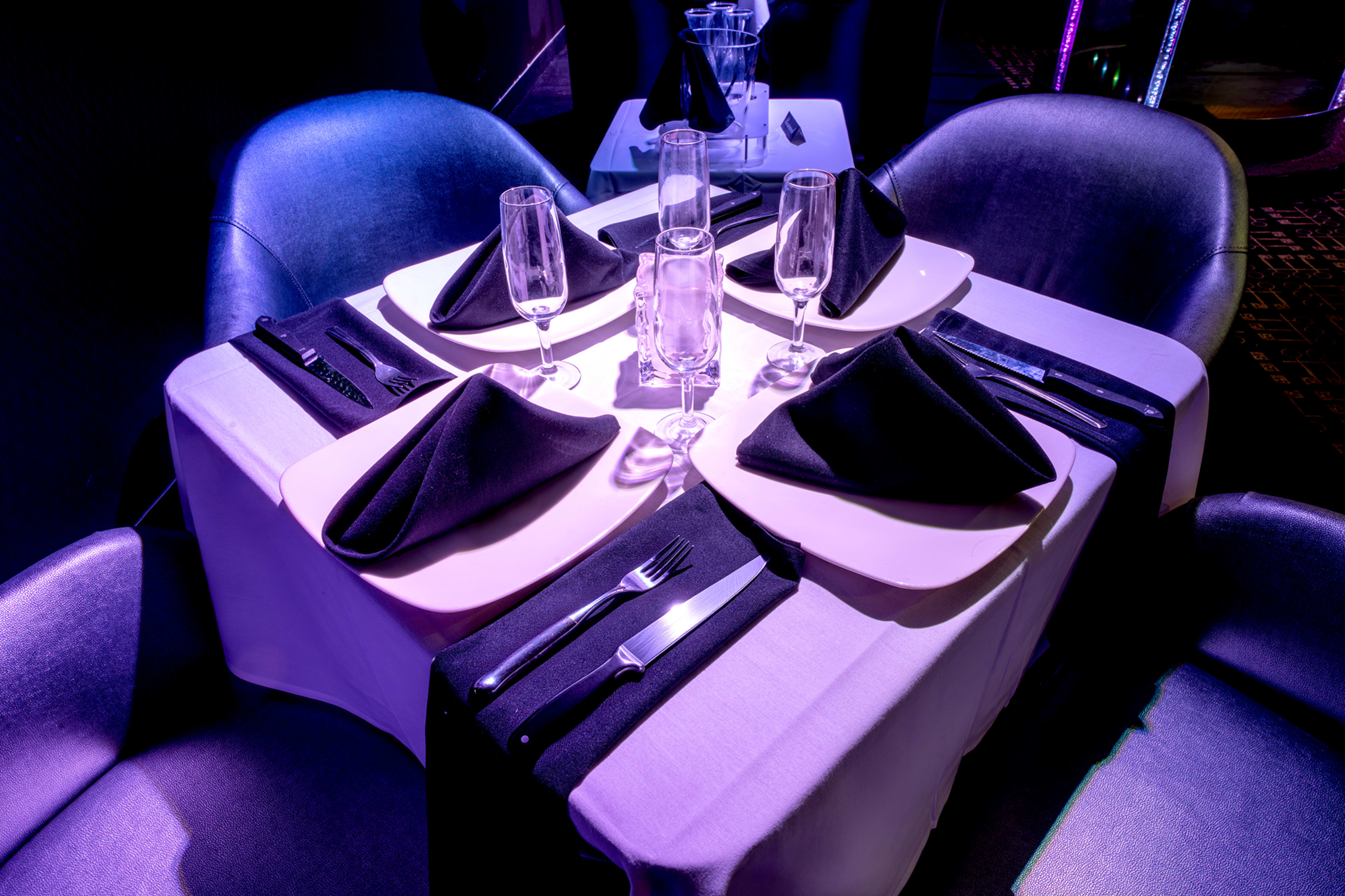 A neatly arranged dining table for four is set with black napkins, glassware, and silverware under soft purple lighting.