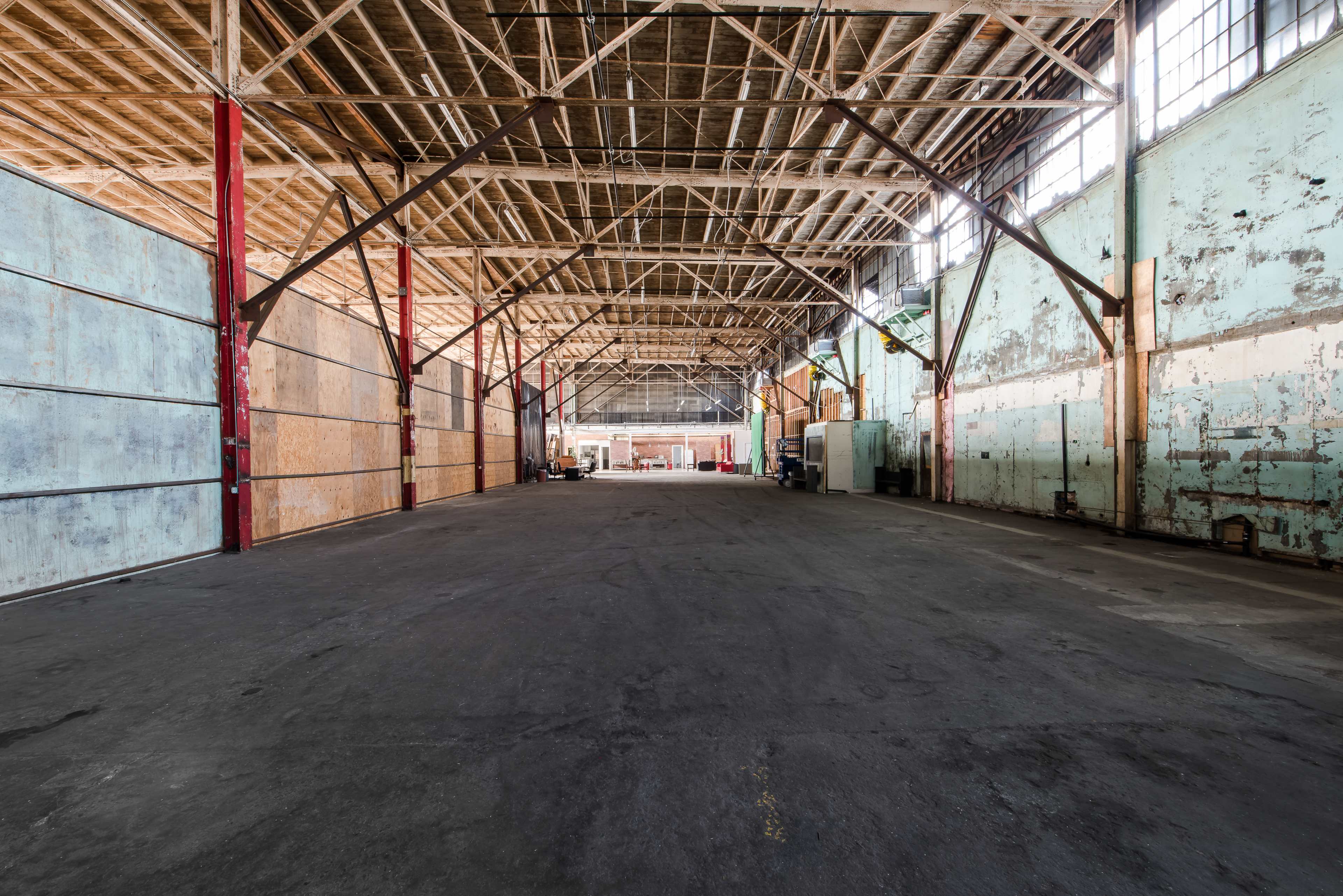 The image shows an empty industrial warehouse with high wooden beams and concrete flooring.