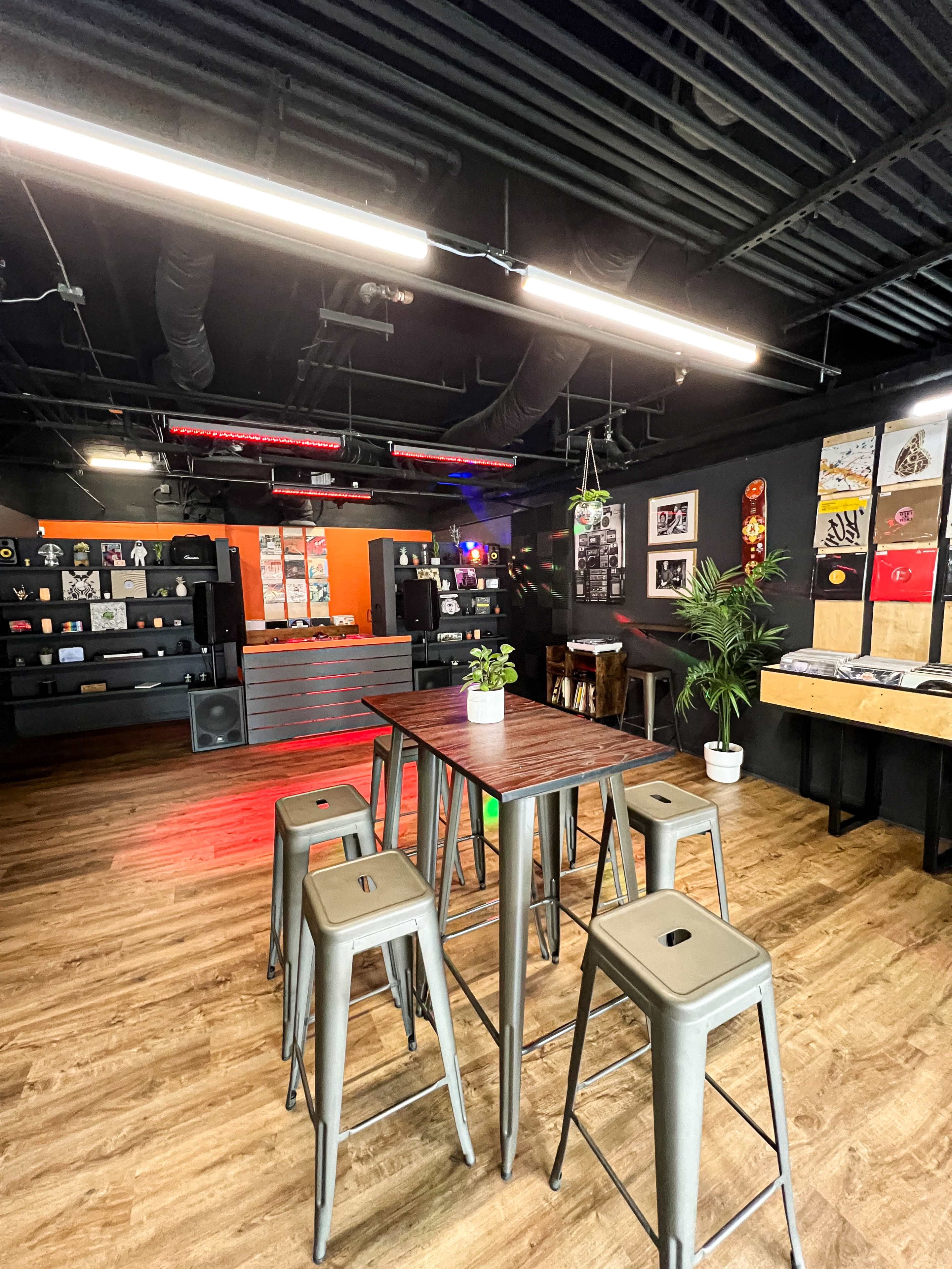 A modern lounge area with a high table surrounded by metal stools, wooden flooring, a wall displaying various artworks, and audio equipment in the background.