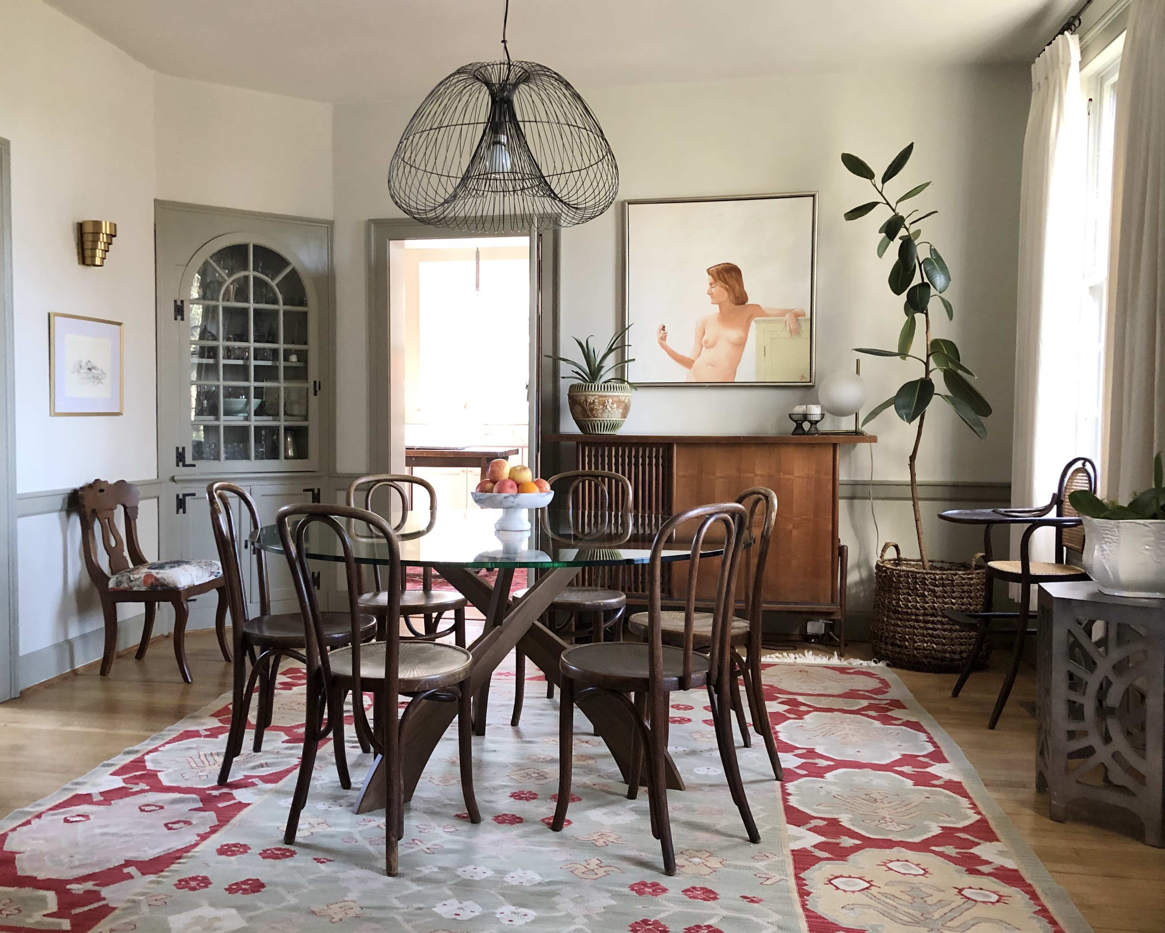 A dining area features a round table with chairs, a large artistic wall piece, and plants beside a sideboard in a well-lit room.