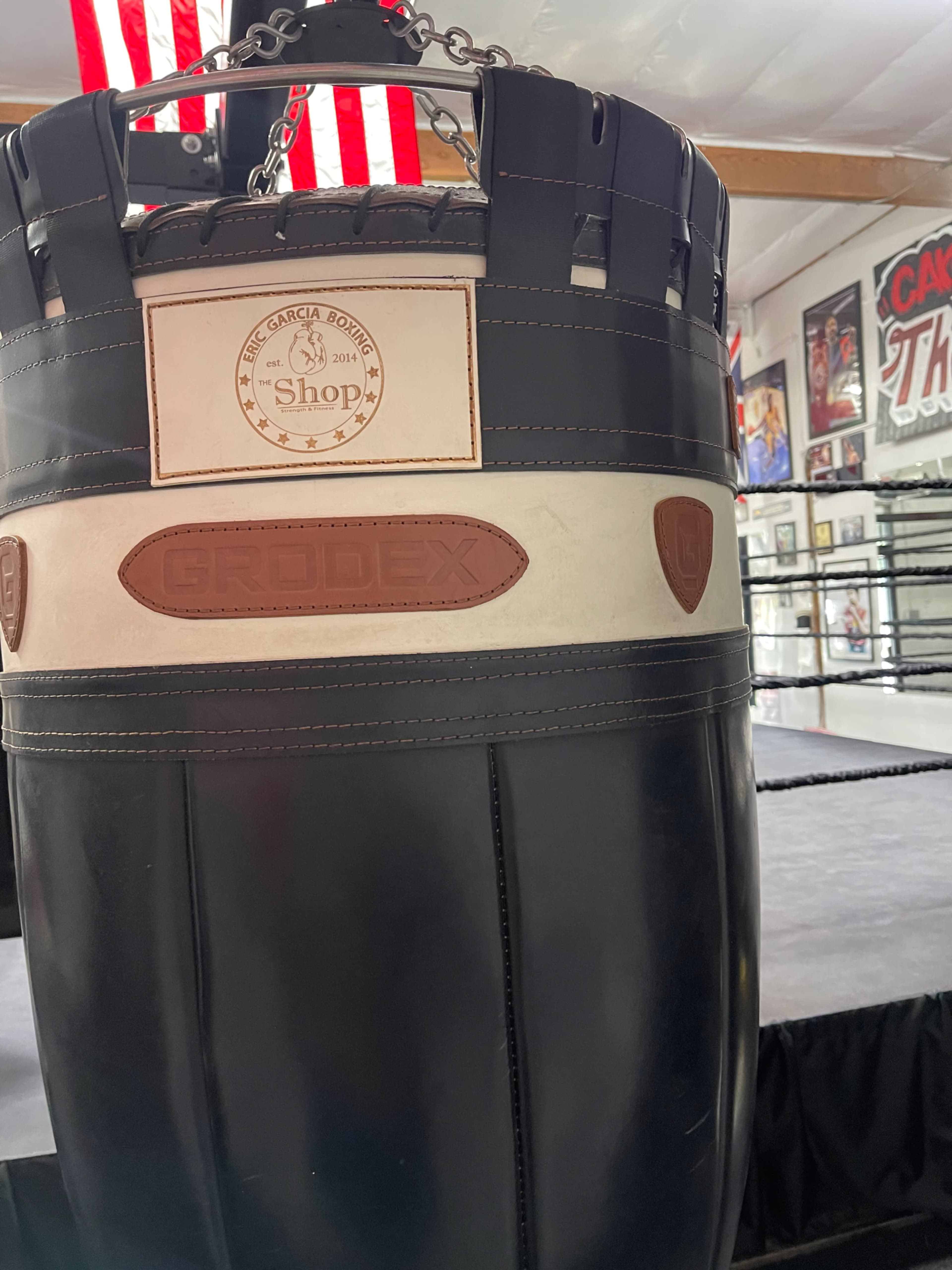 A black punching bag with a logo in a boxing gym, featuring a ring and American flags in the background.