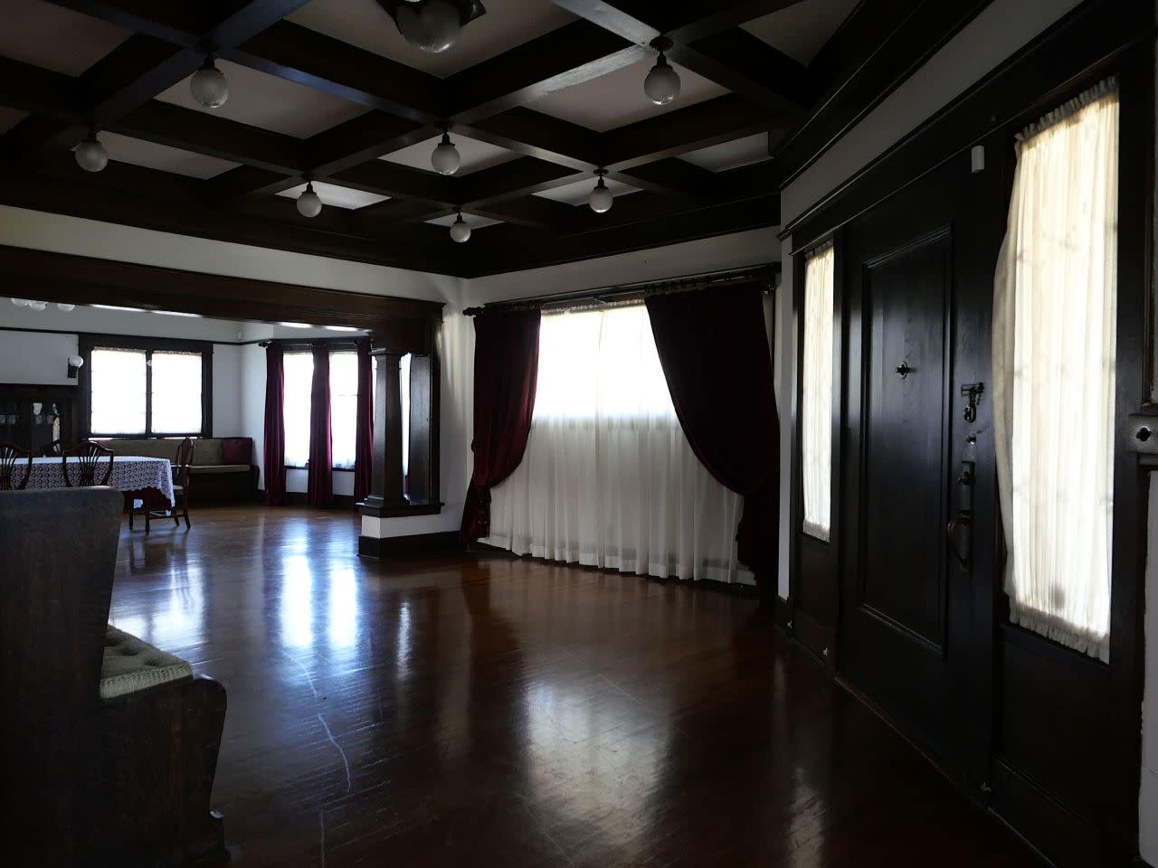 The image shows a spacious room with wooden beams, large windows draped in white and burgundy curtains, and polished wooden flooring.