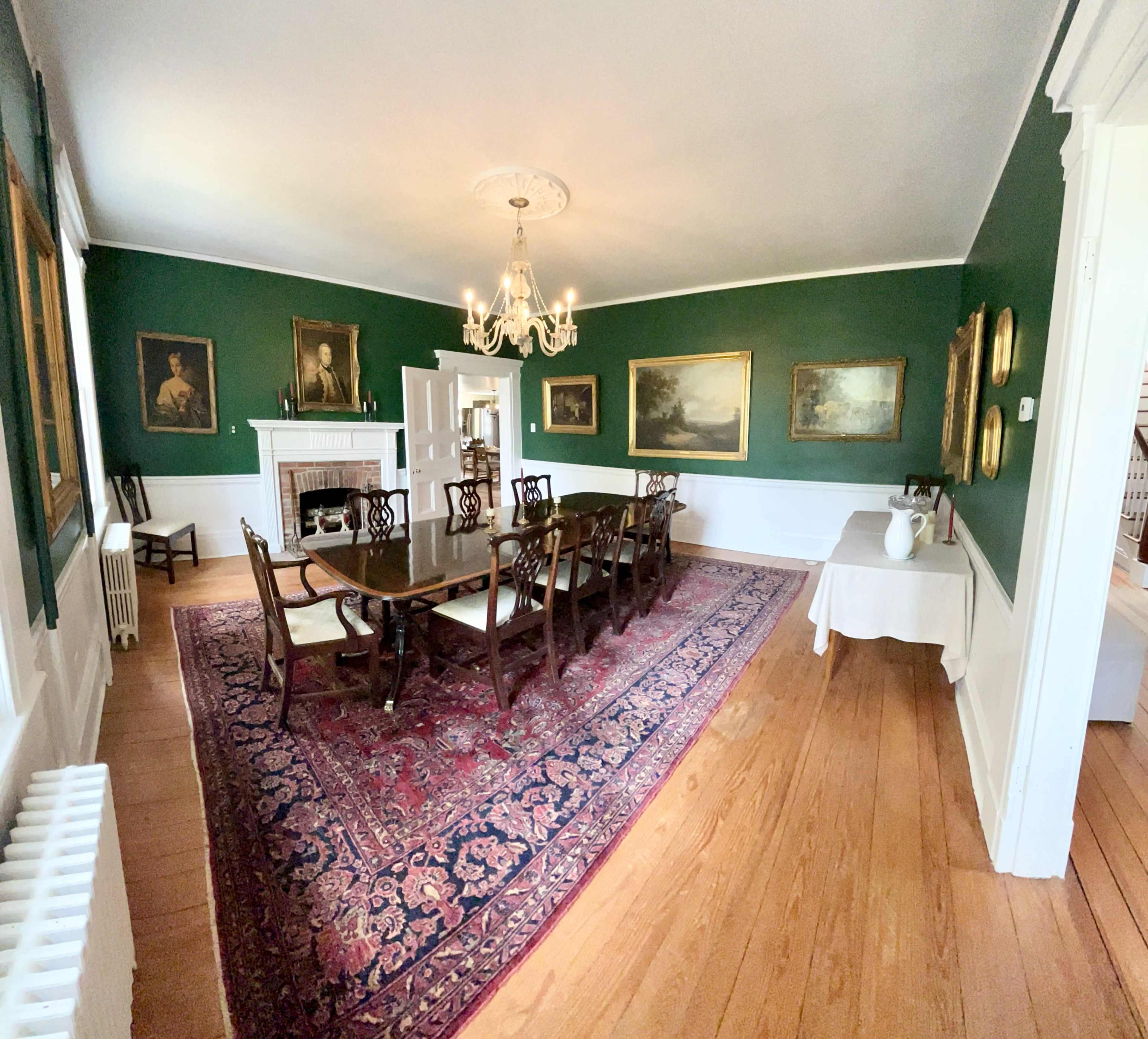 A dining room with a large wooden table, surrounded by chairs, set against green walls adorned with framed paintings and a chandelier overhead.
