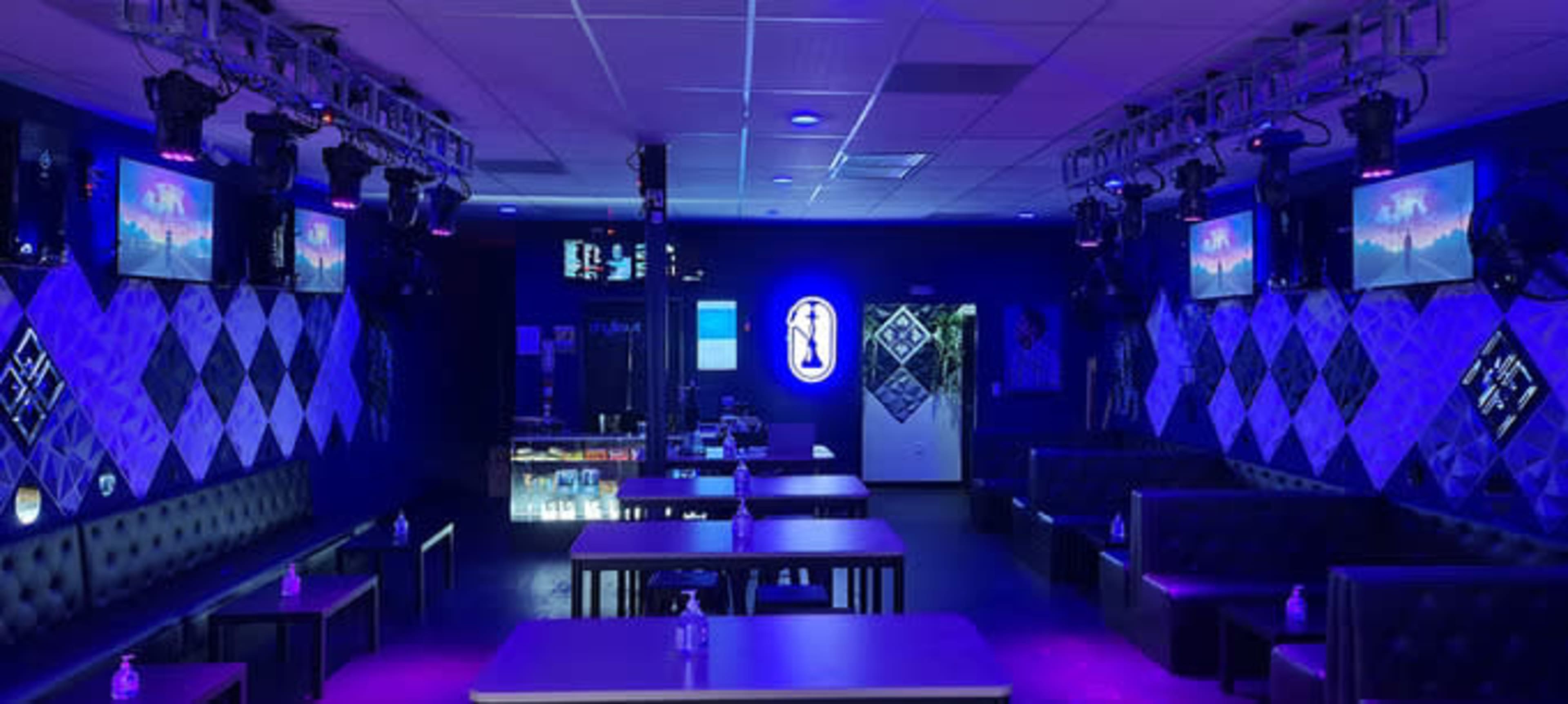 The image shows a dimly lit lounge with purple lighting, featuring several tables and wall-mounted screens.