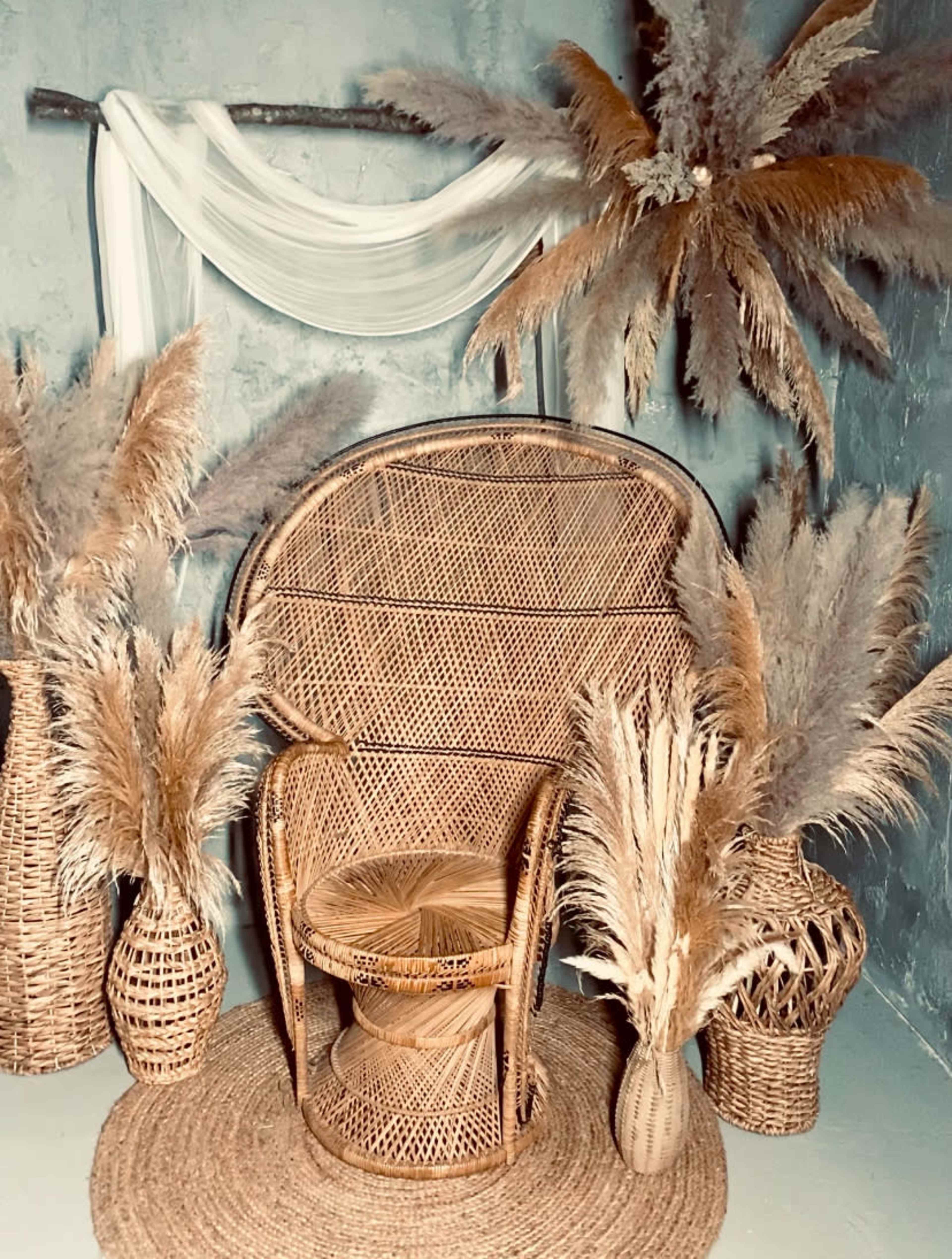 A bohemian-inspired seating arrangement with a rattan chair, a small stool, and pampas grass in woven baskets, set against a textured blue background.