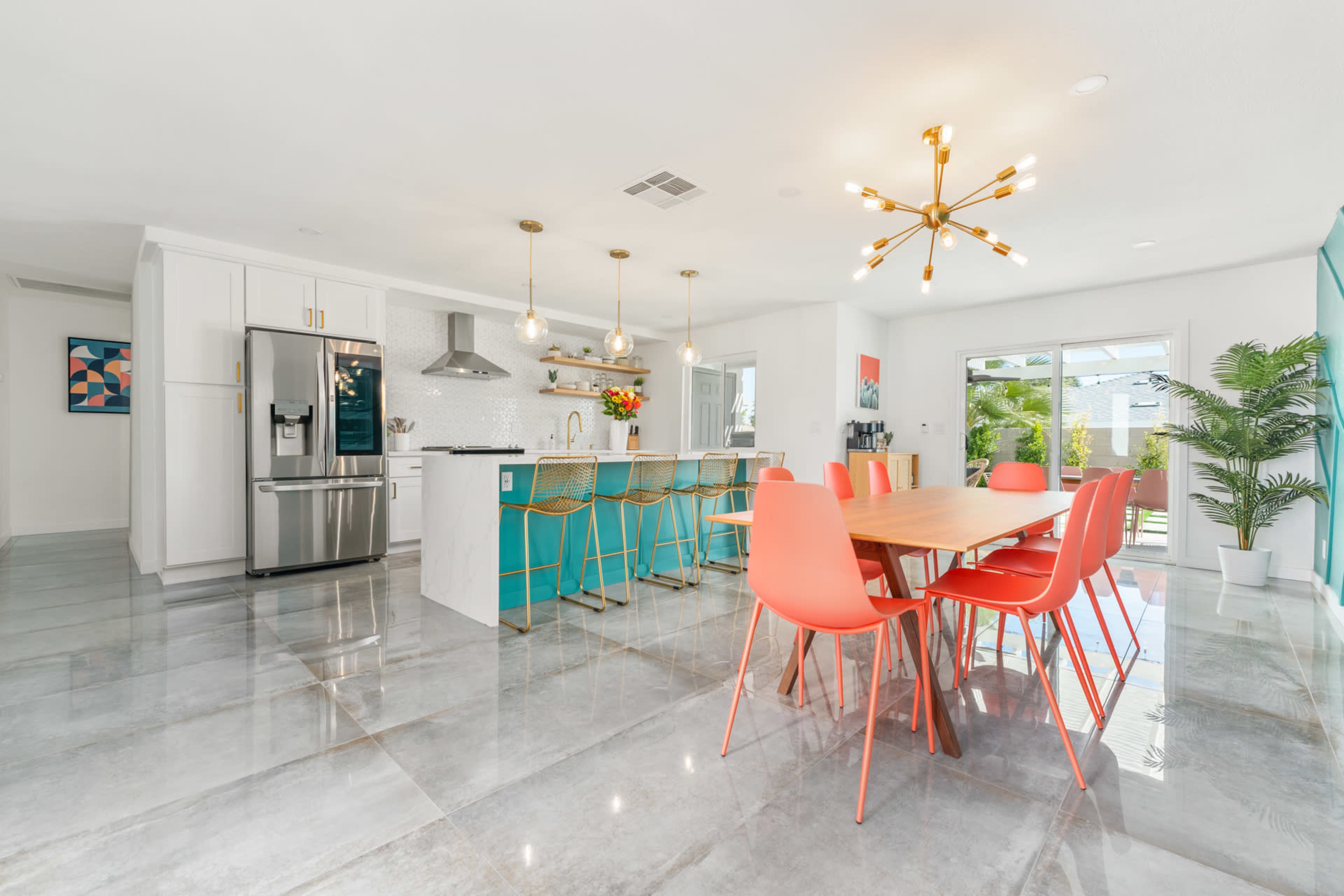 A modern kitchen and dining area feature an island with turquoise cabinetry, stainless steel appliances, and a long wooden table surrounded by bright pink chairs.