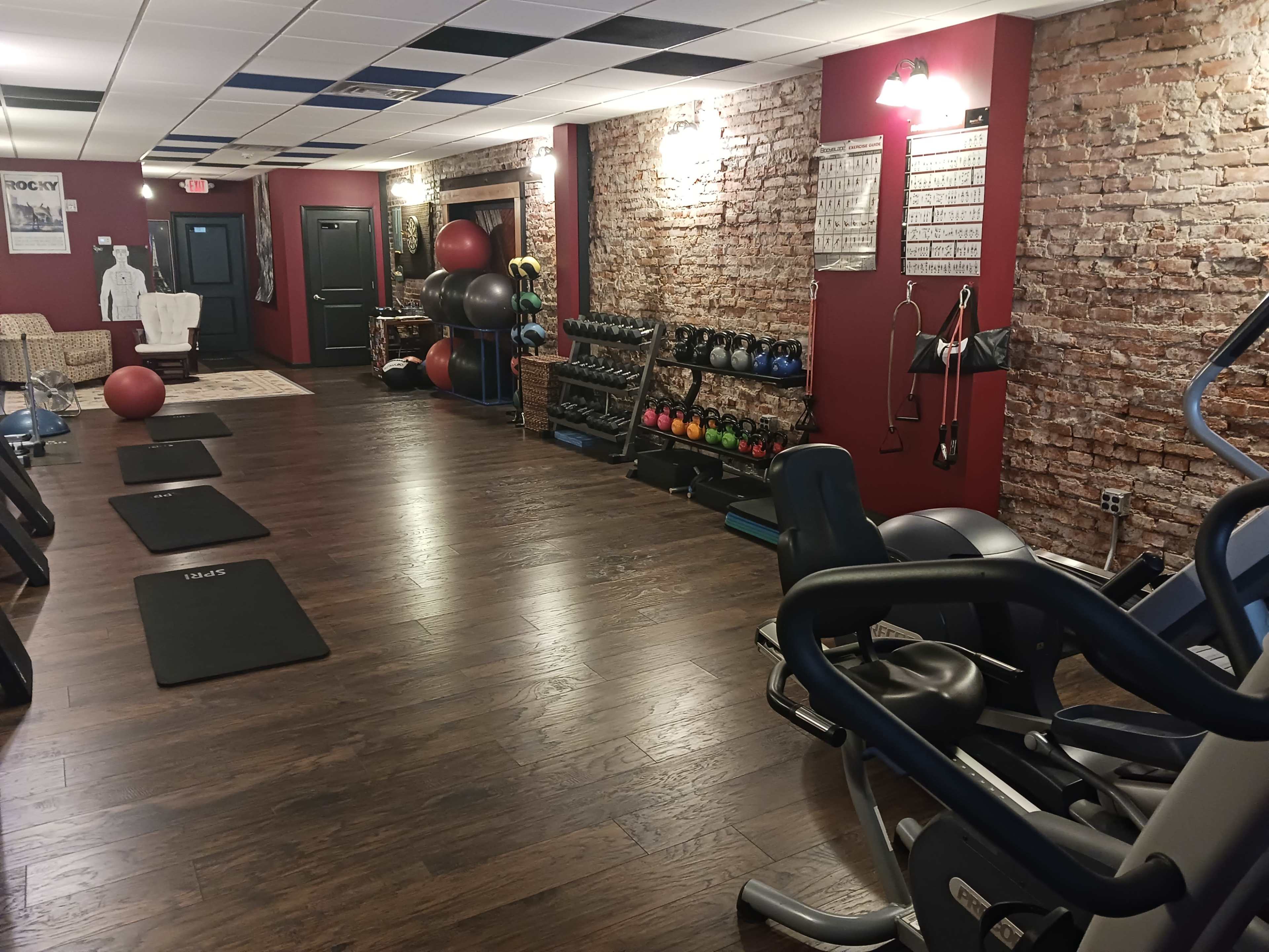 The image shows a well-equipped fitness area with exercise mats, various weights, and workout machines against a brick wall.