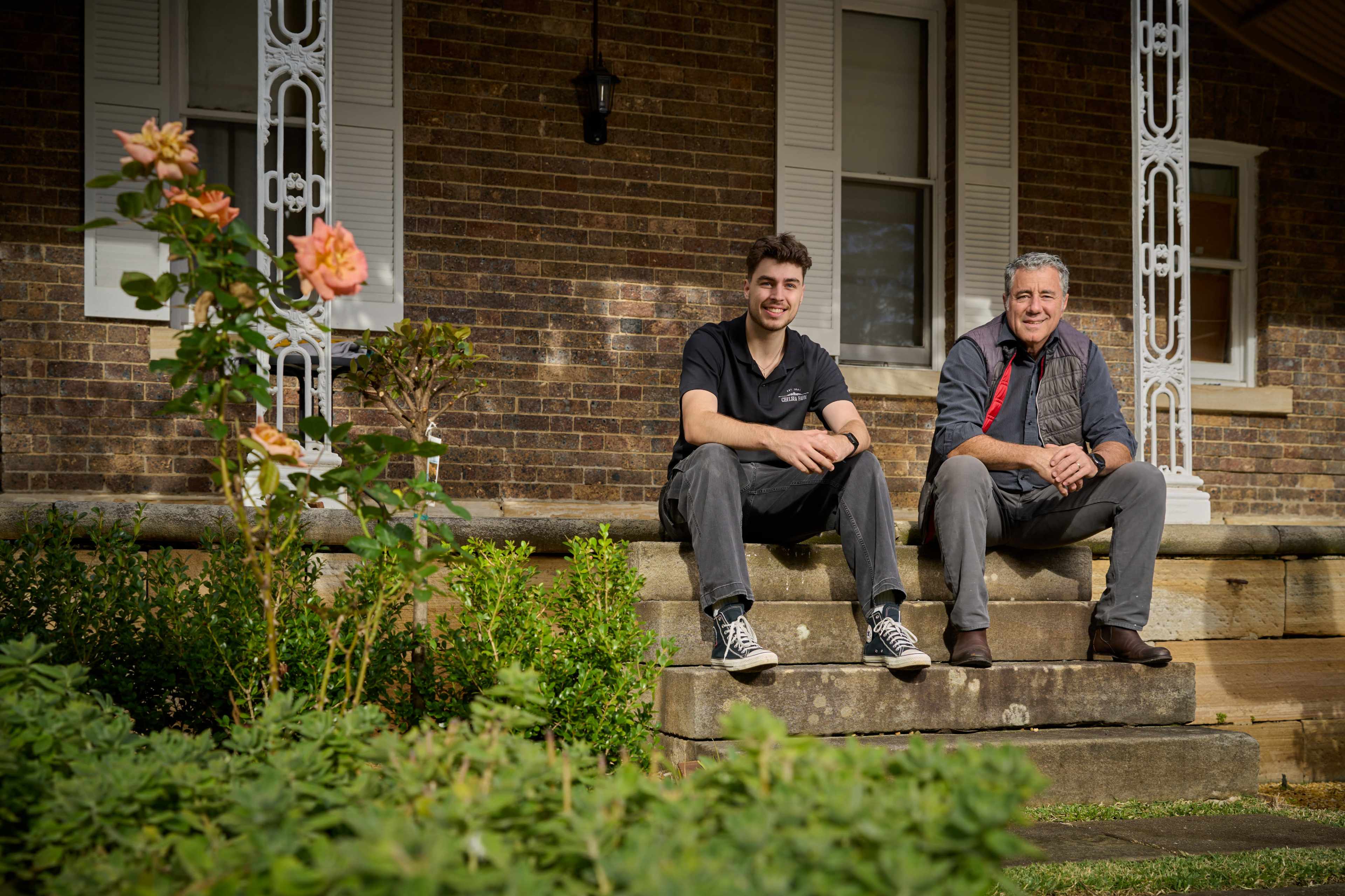 Two men sit on the steps of a brick building, with a rose bush in bloom nearby.