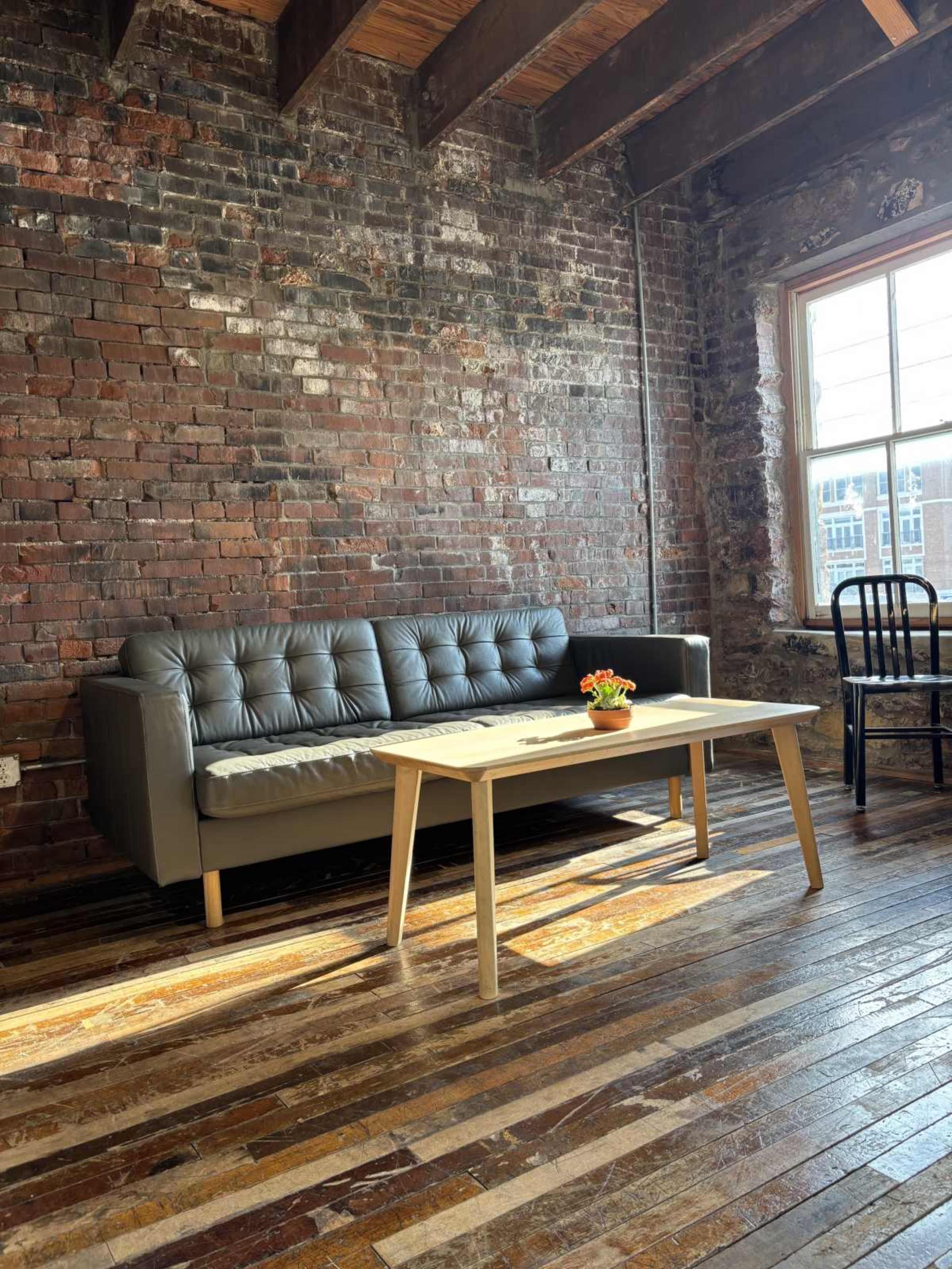 The scene features a modern gray couch and a wooden coffee table positioned on a hardwood floor in a room with exposed brick walls and large windows.