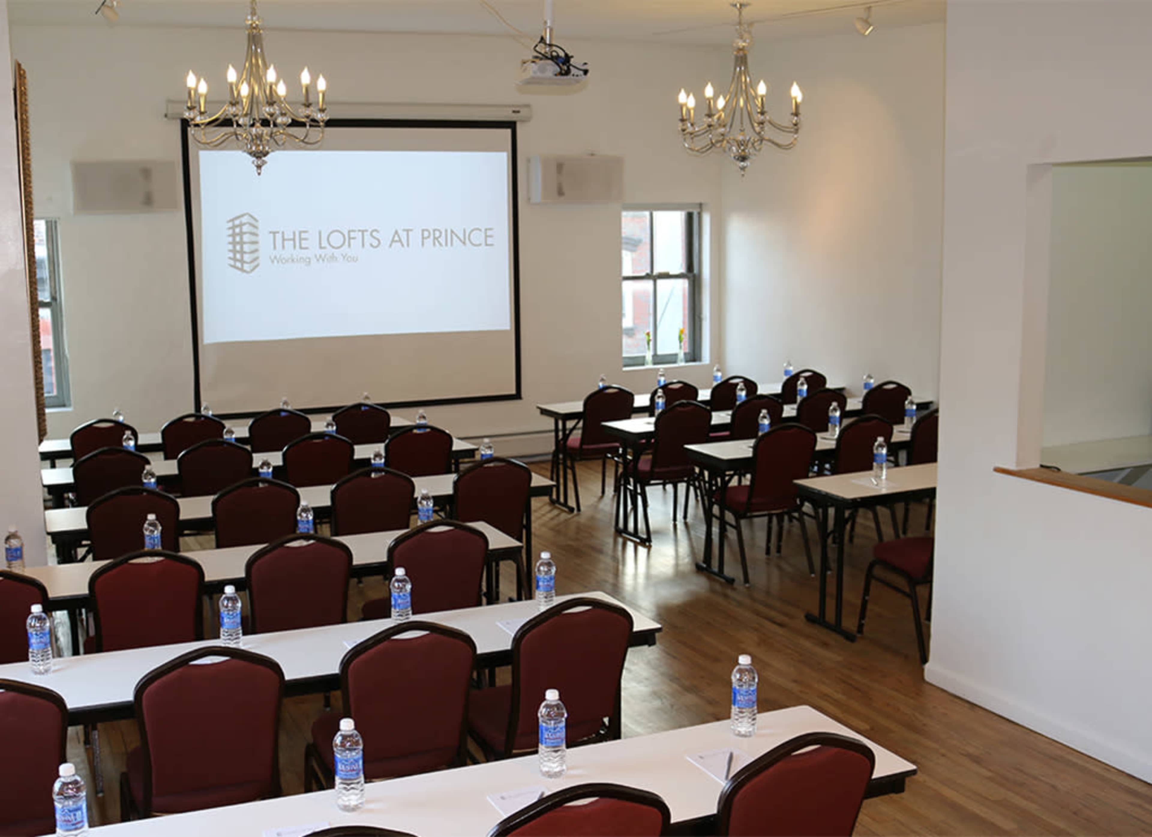 The image shows a conference room set up with rows of tables and chairs, each equipped with bottles of water, facing a projector screen displaying a logo.