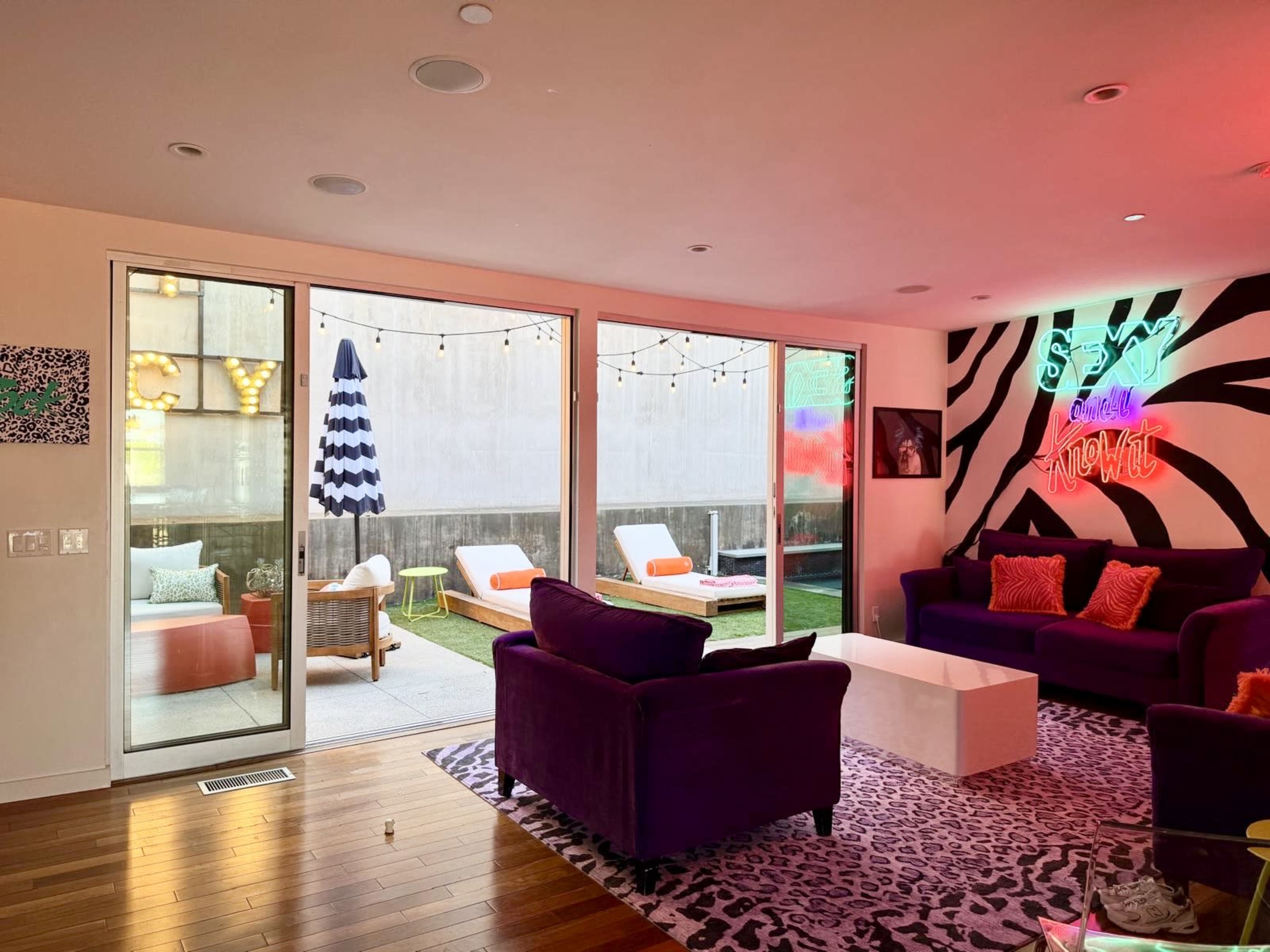 The room features a vibrant seating area with purple couches and a white coffee table, alongside large glass doors leading to an outdoor space with colorful lounge furniture and string lights.