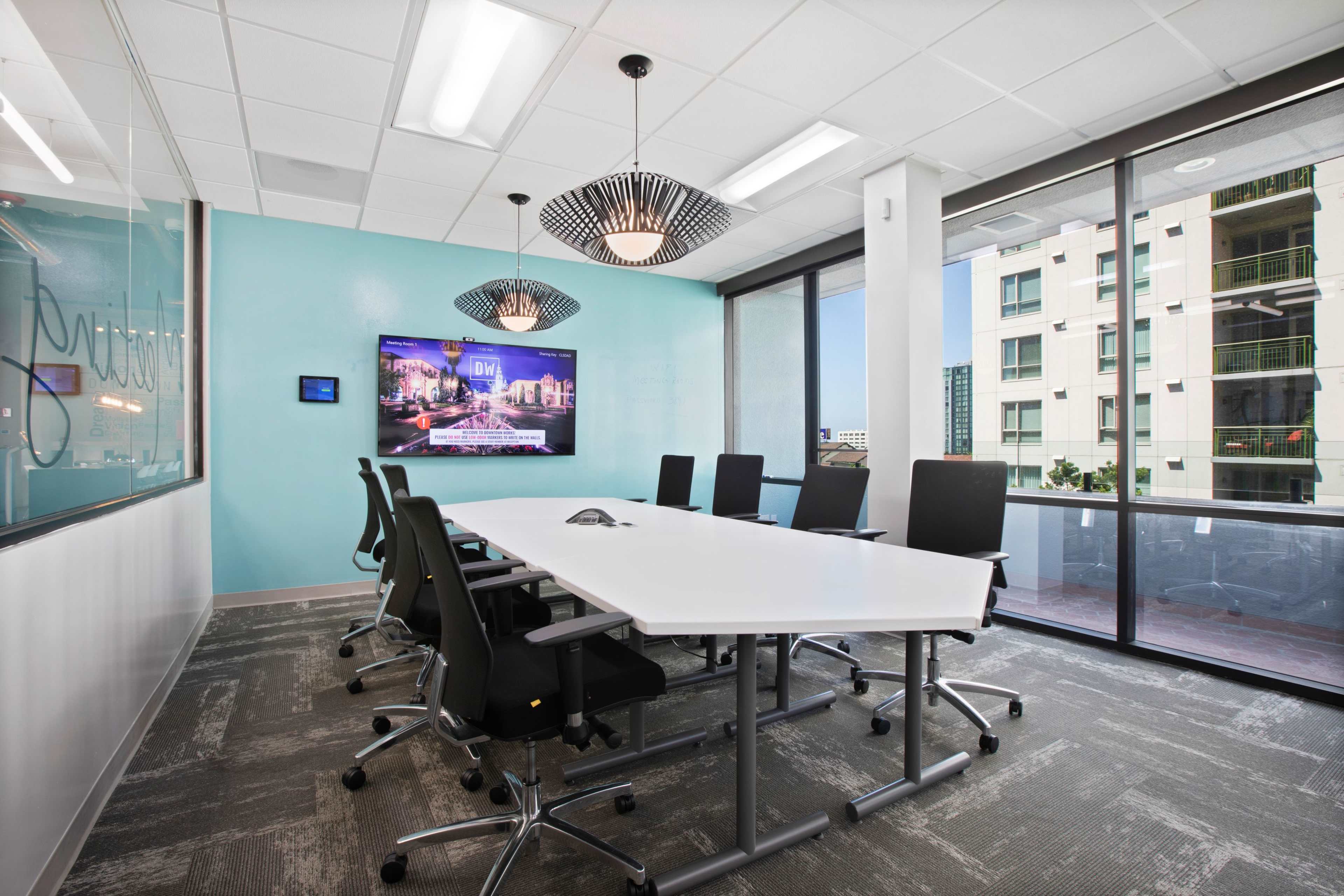 A modern conference room features a large table surrounded by black chairs, a wall-mounted screen displaying a cityscape, and large windows overlooking an outdoor area.