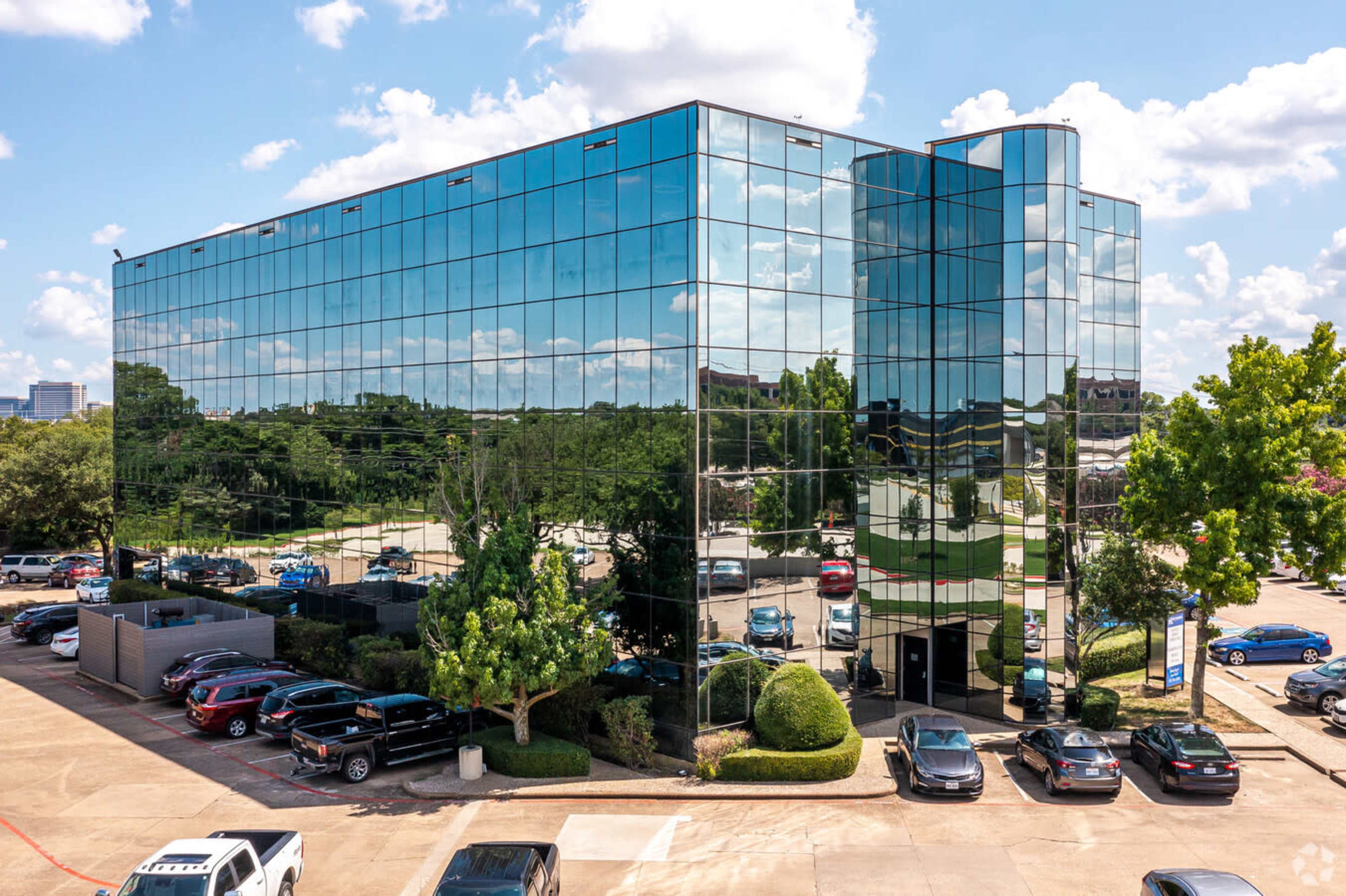 A modern glass office building with reflective windows stands among parked cars and trees under a blue sky with clouds.