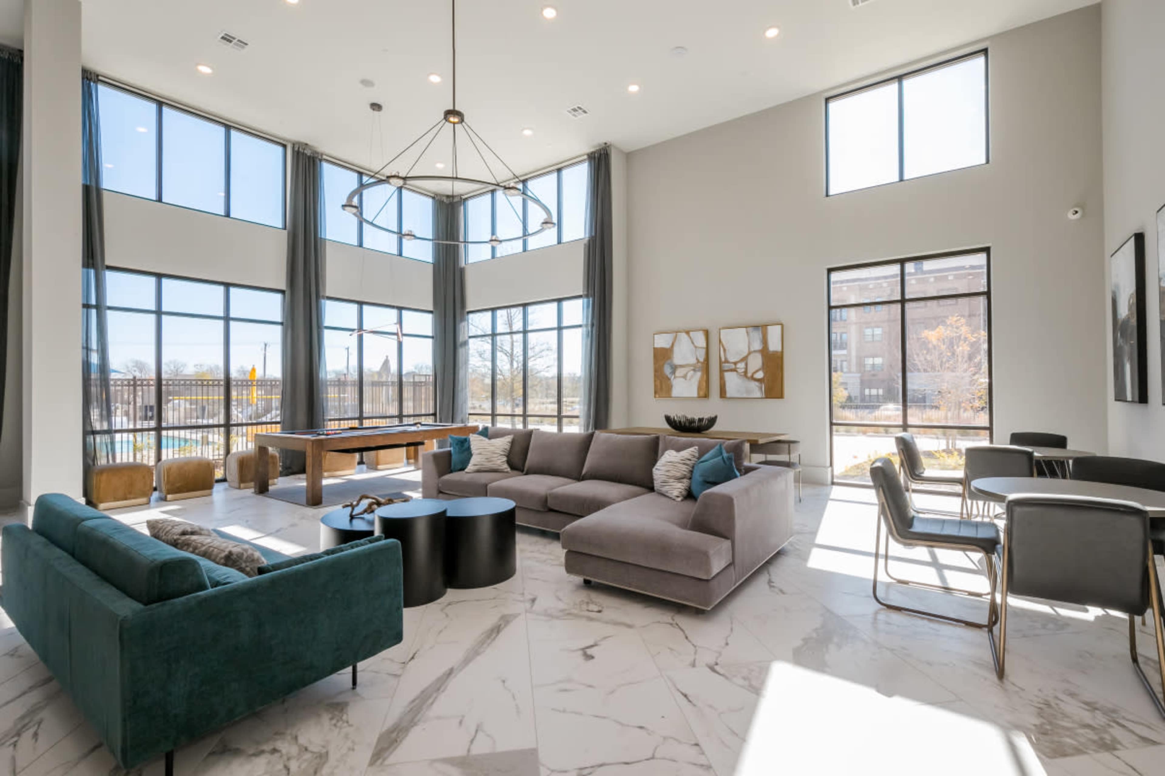 The image shows a spacious, modern living area with large windows, a sectional couch, and a dining area featuring tables and chairs.