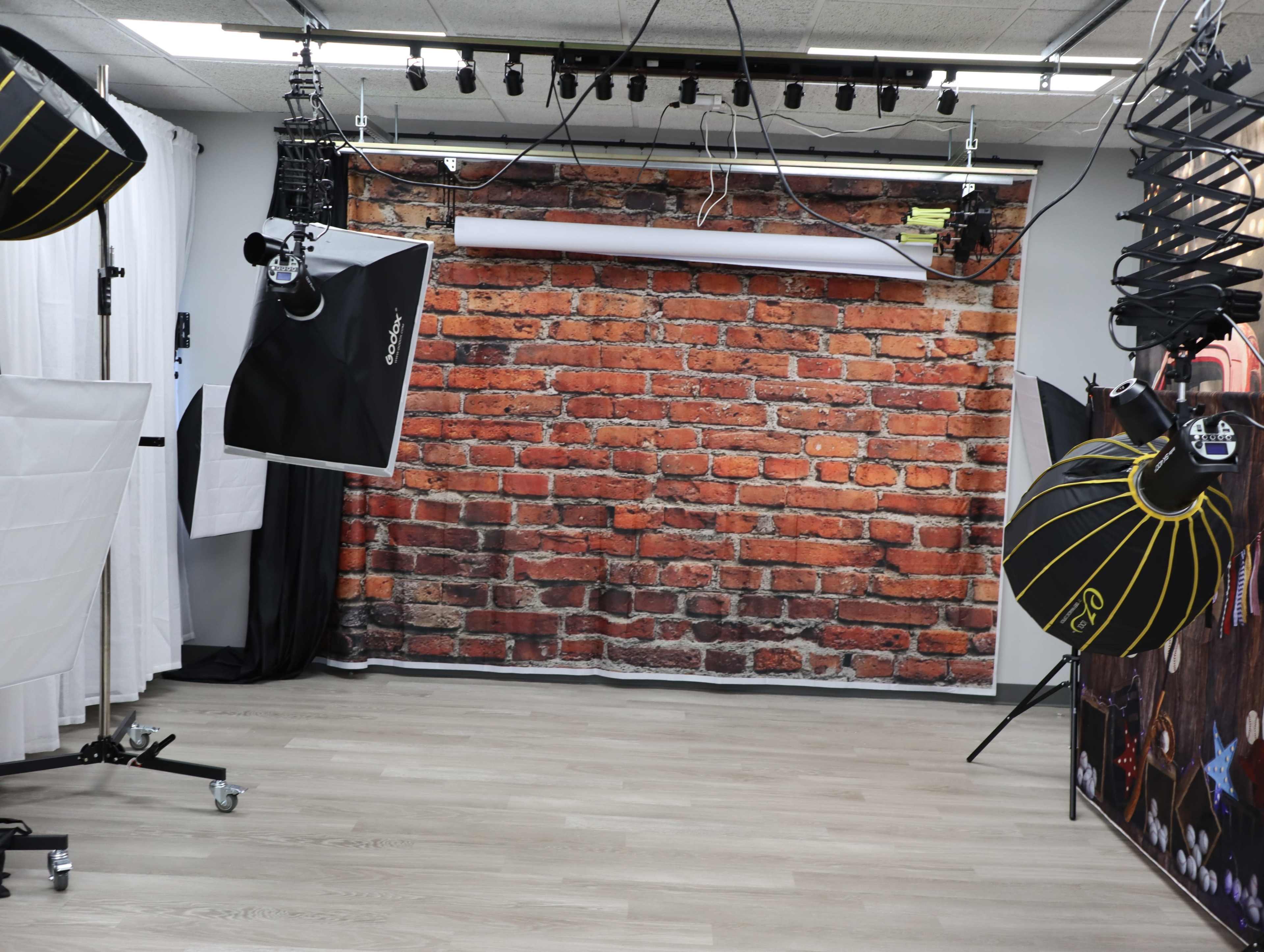 The image shows a photography studio with a brick wall backdrop, various lighting equipment, and a wooden floor.