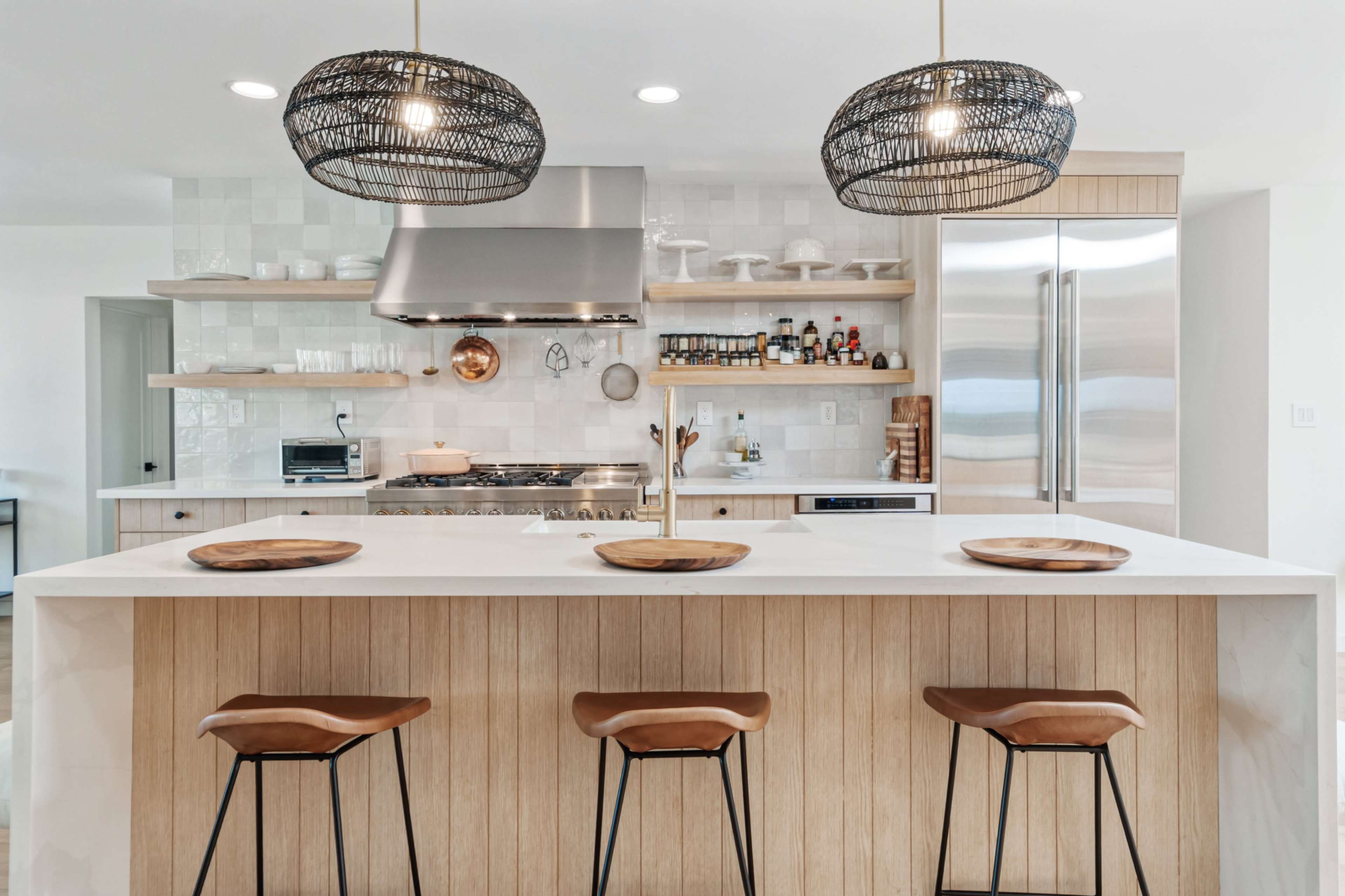 The image shows a modern kitchen with a central island featuring three wooden bar stools, pendant lights hanging above, a stainless-steel range hood, and open shelves displaying kitchenware.