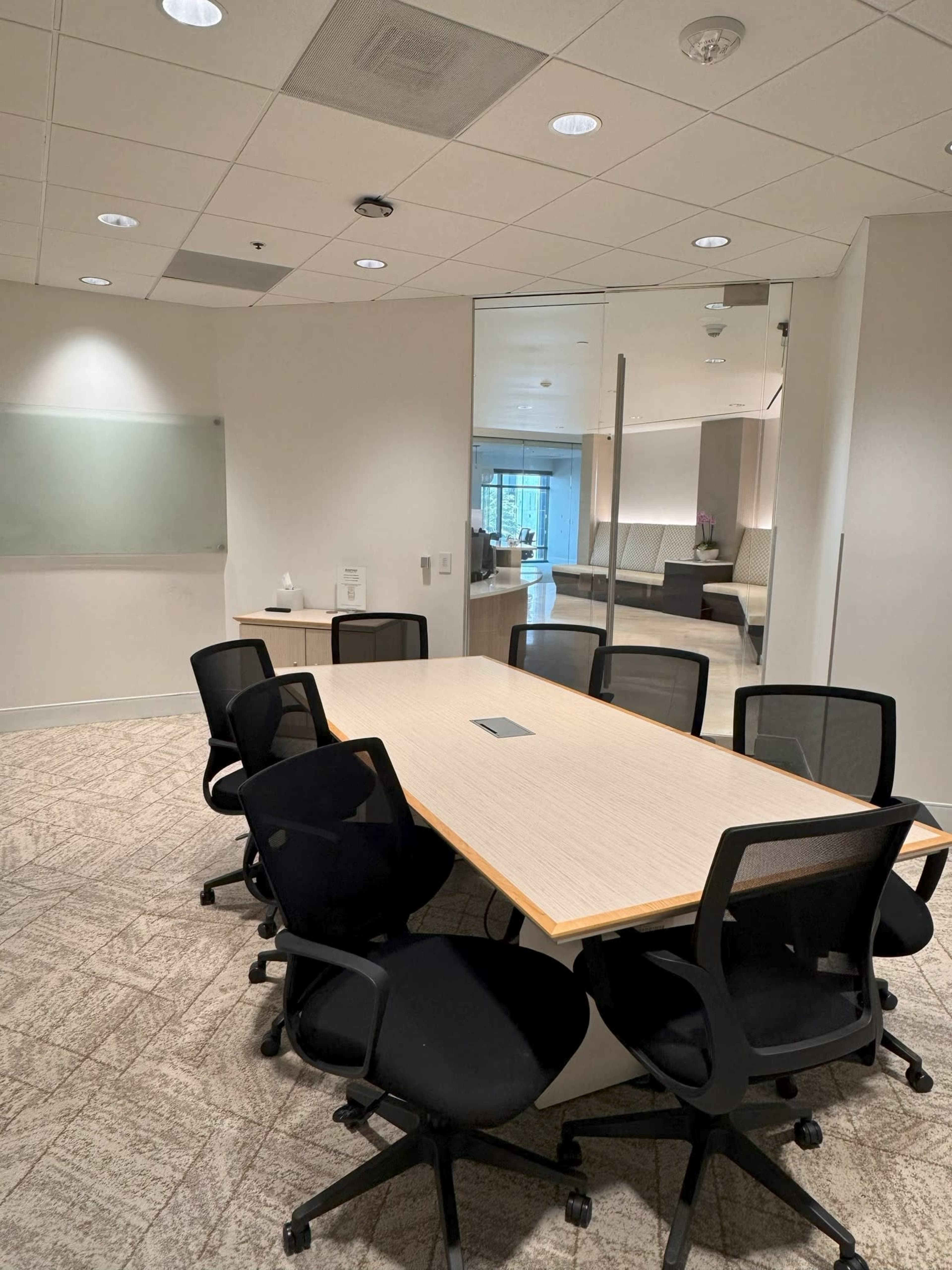 The image shows a modern conference room with a large table surrounded by several office chairs, and a glass wall leading to a lounge area.