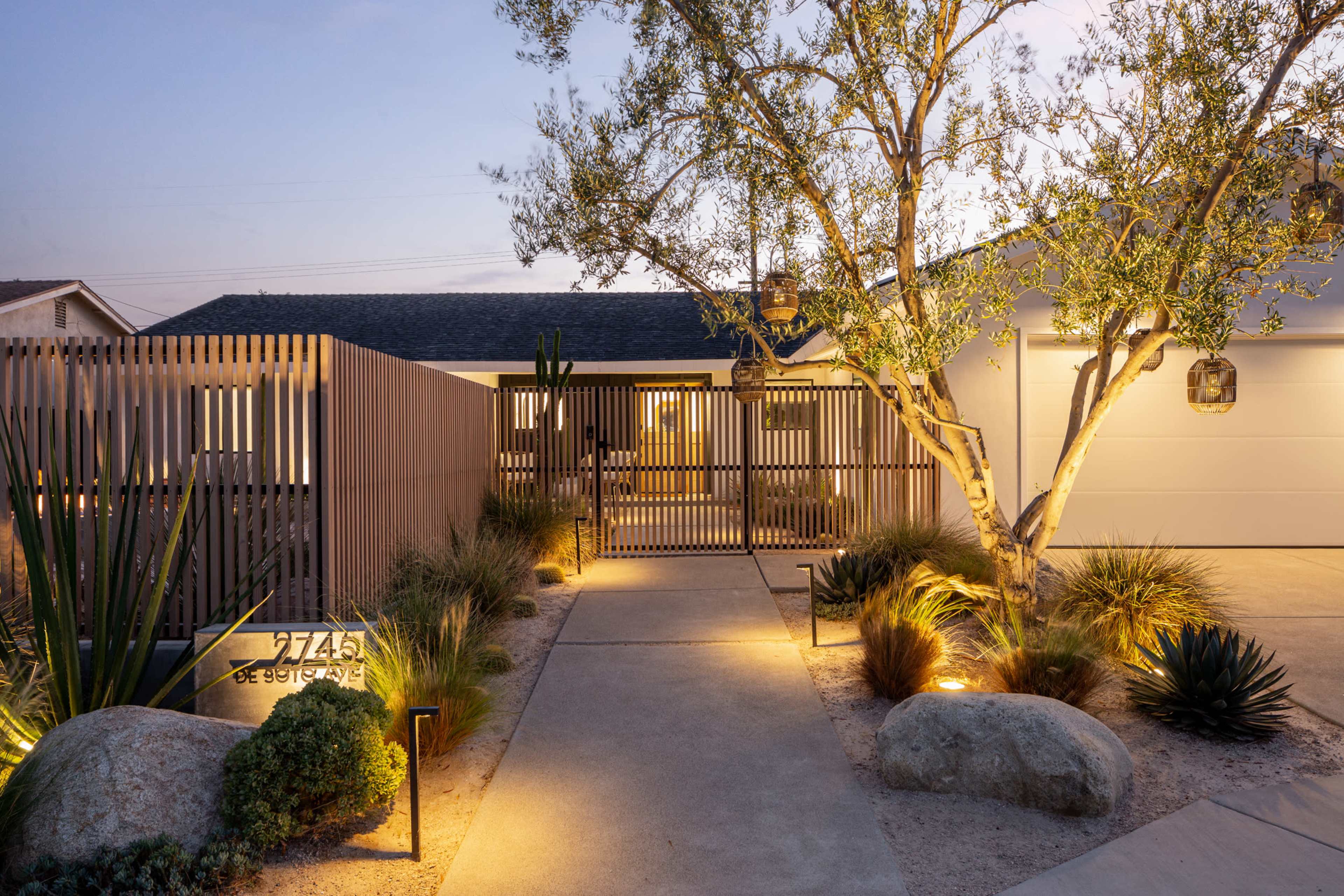 A modern home entrance is highlighted by landscaped paths, decorative lighting, and a wooden gate surrounded by plants and large rocks.