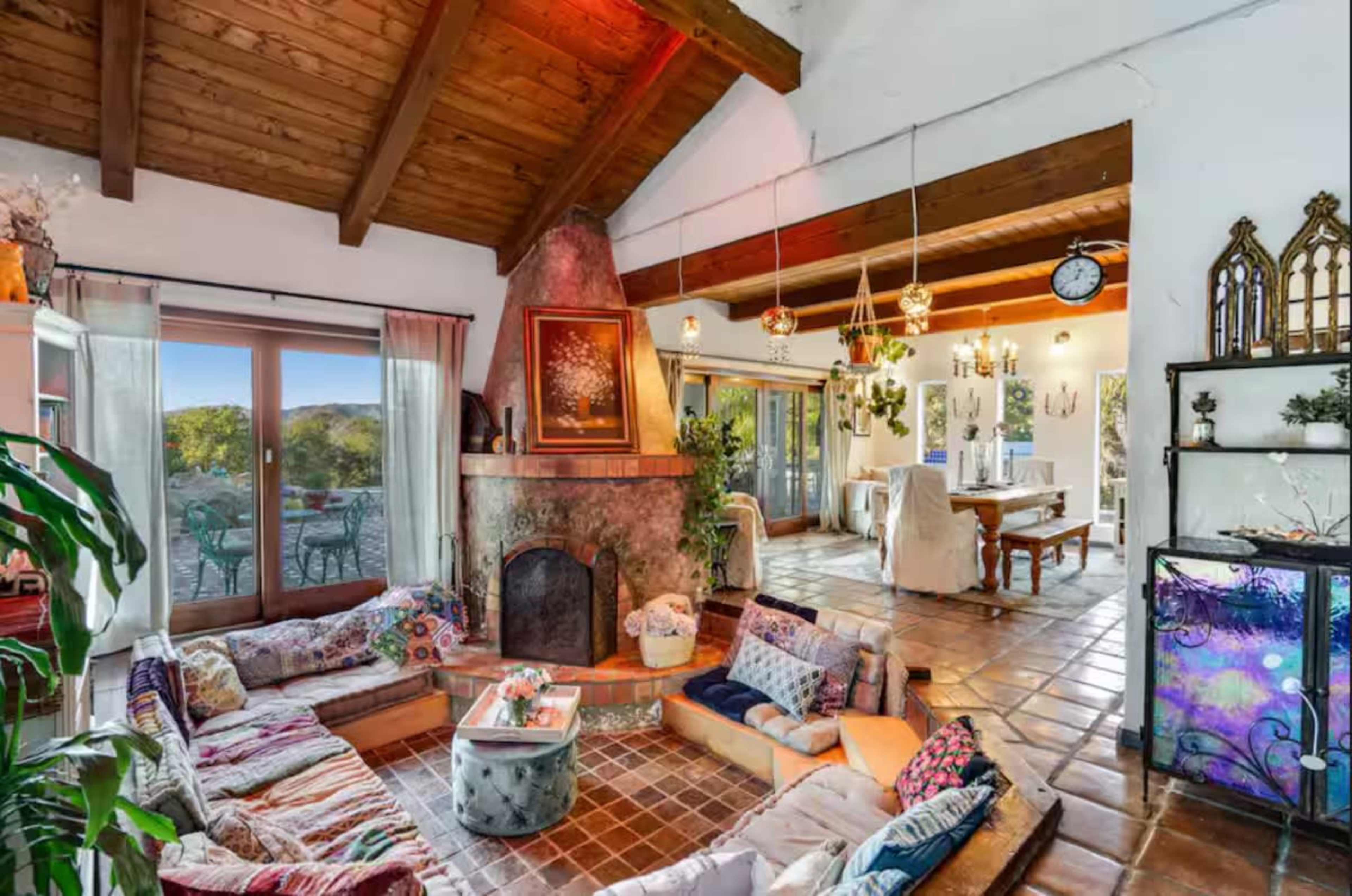The image shows a cozy living room with a large stone fireplace, wooden beams on the ceiling, and a seating area arranged with colorful cushions surrounding a central table.