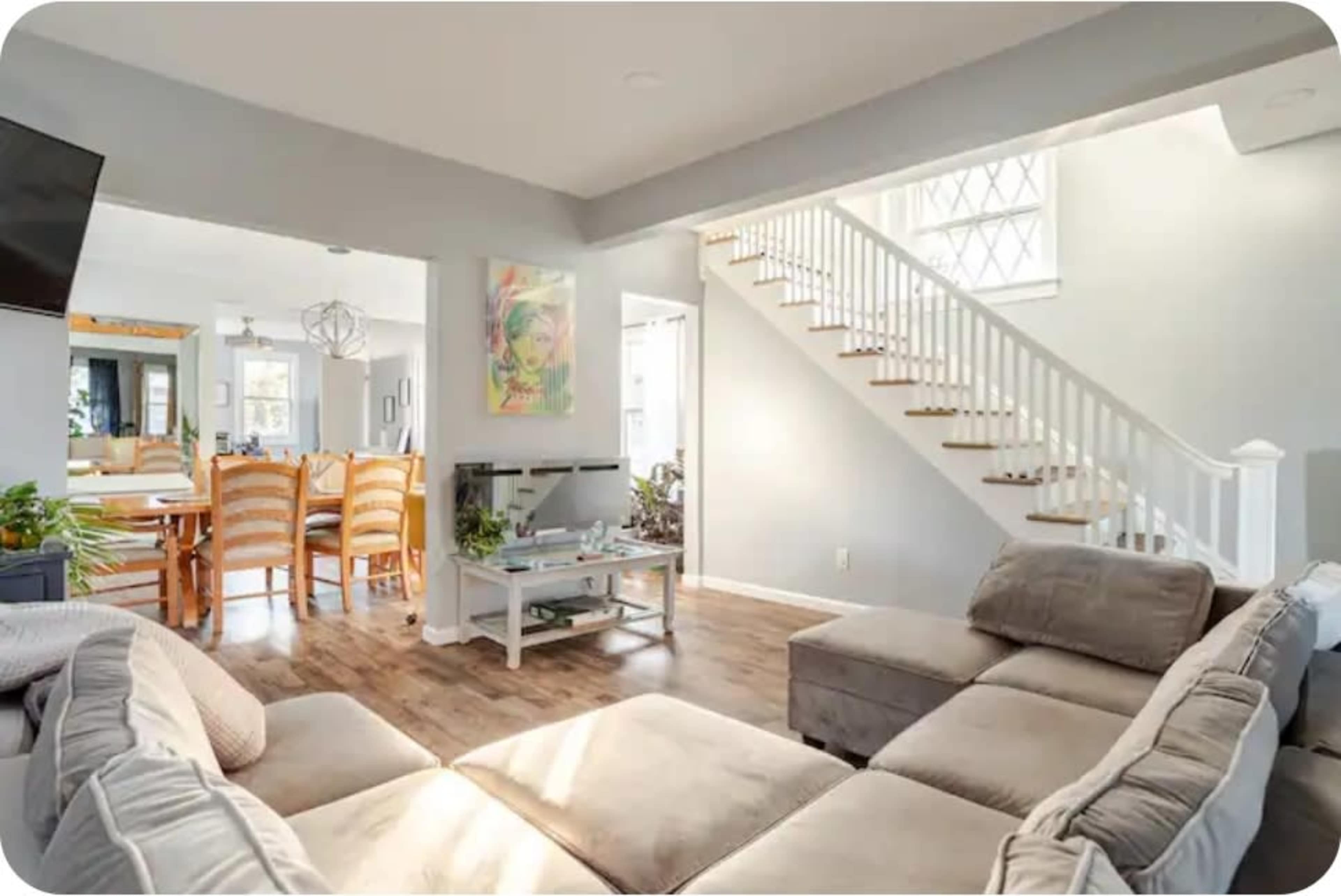 A modern living room features a sectional sofa, a coffee table, and a staircase leading to an upper level, with a dining area visible through an archway.
