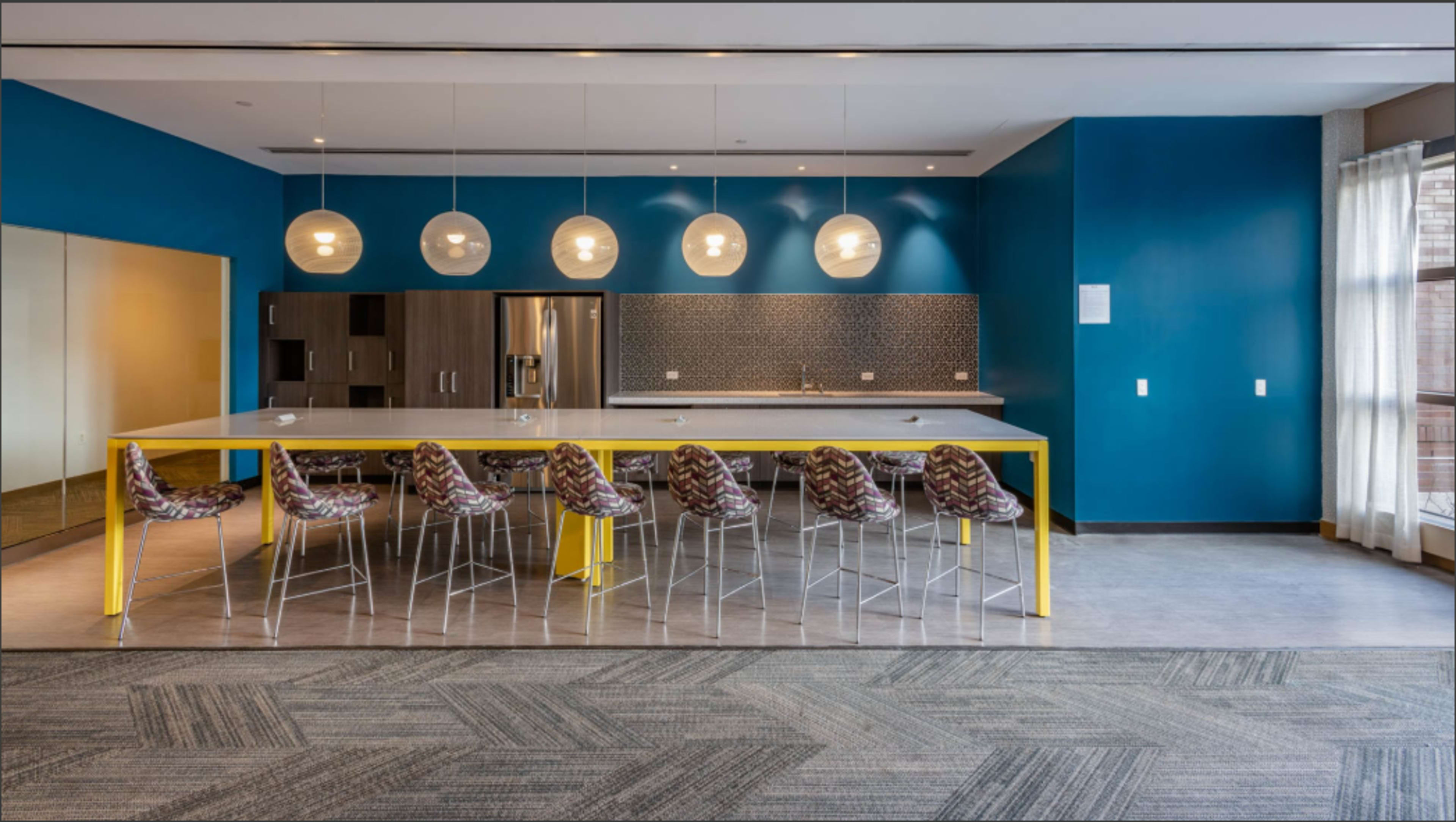A modern kitchen area with a long white table supported by a yellow base, surrounded by stylish bar stools, and featuring pendant lighting above.