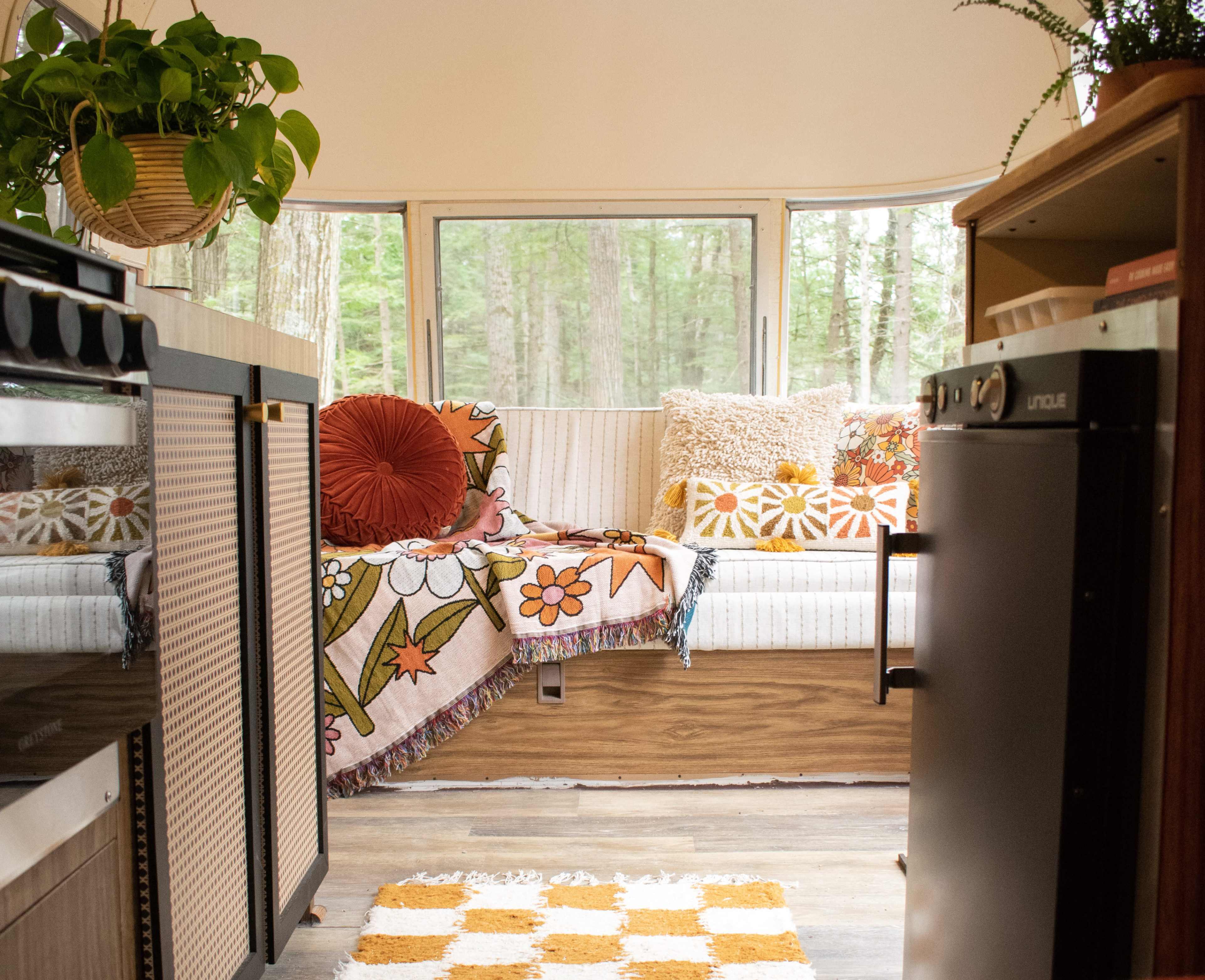 An interior of a camper with a cozy seating area, a patterned blanket, and views of greenery through large windows.