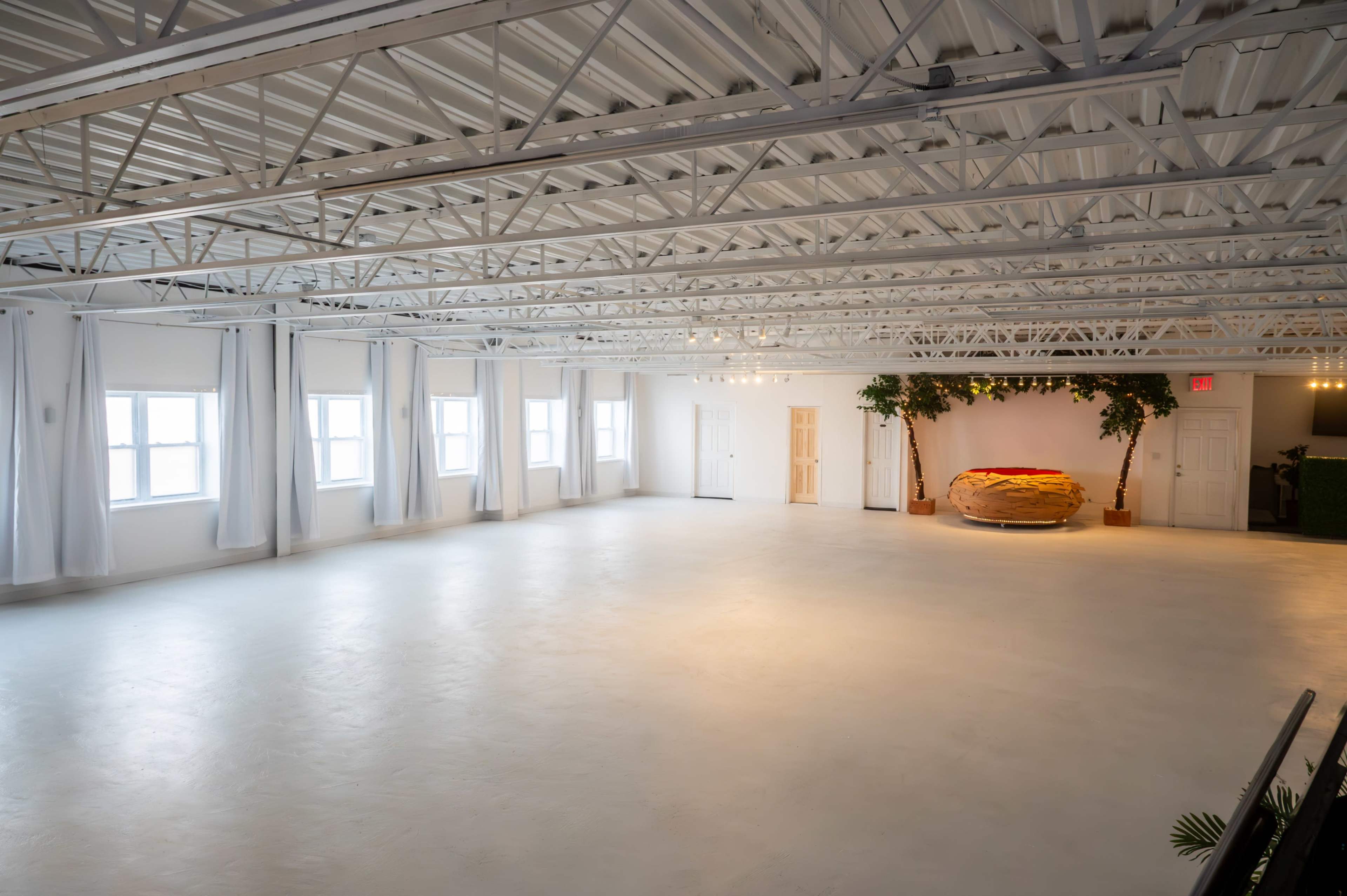 The image shows a spacious, empty room with white walls, large windows, and a decorative area featuring a circular, orange object surrounded by greenery.