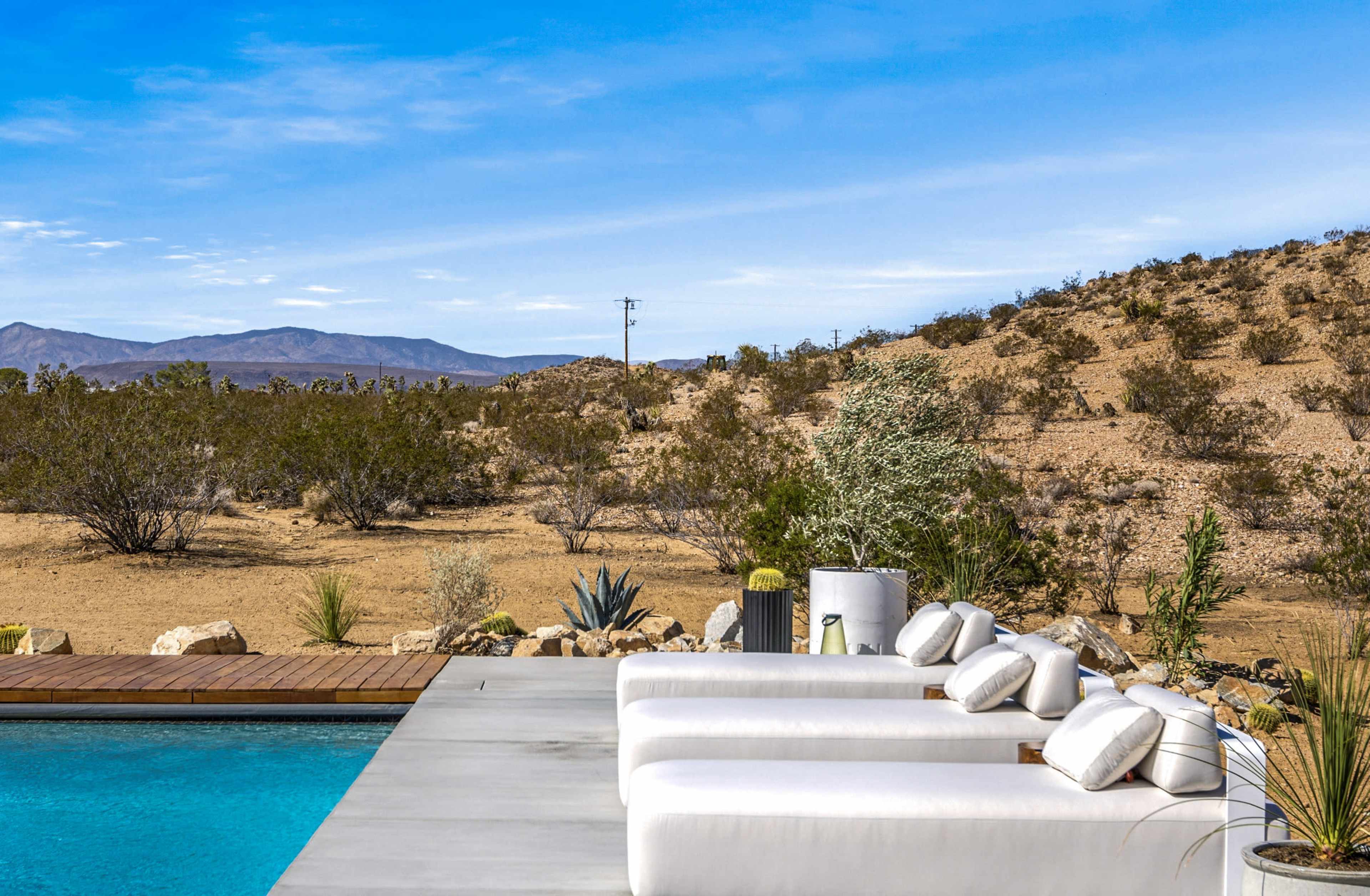 A modern patio with white loungers overlooks a swimming pool and a desert landscape with sparse vegetation and distant mountains.