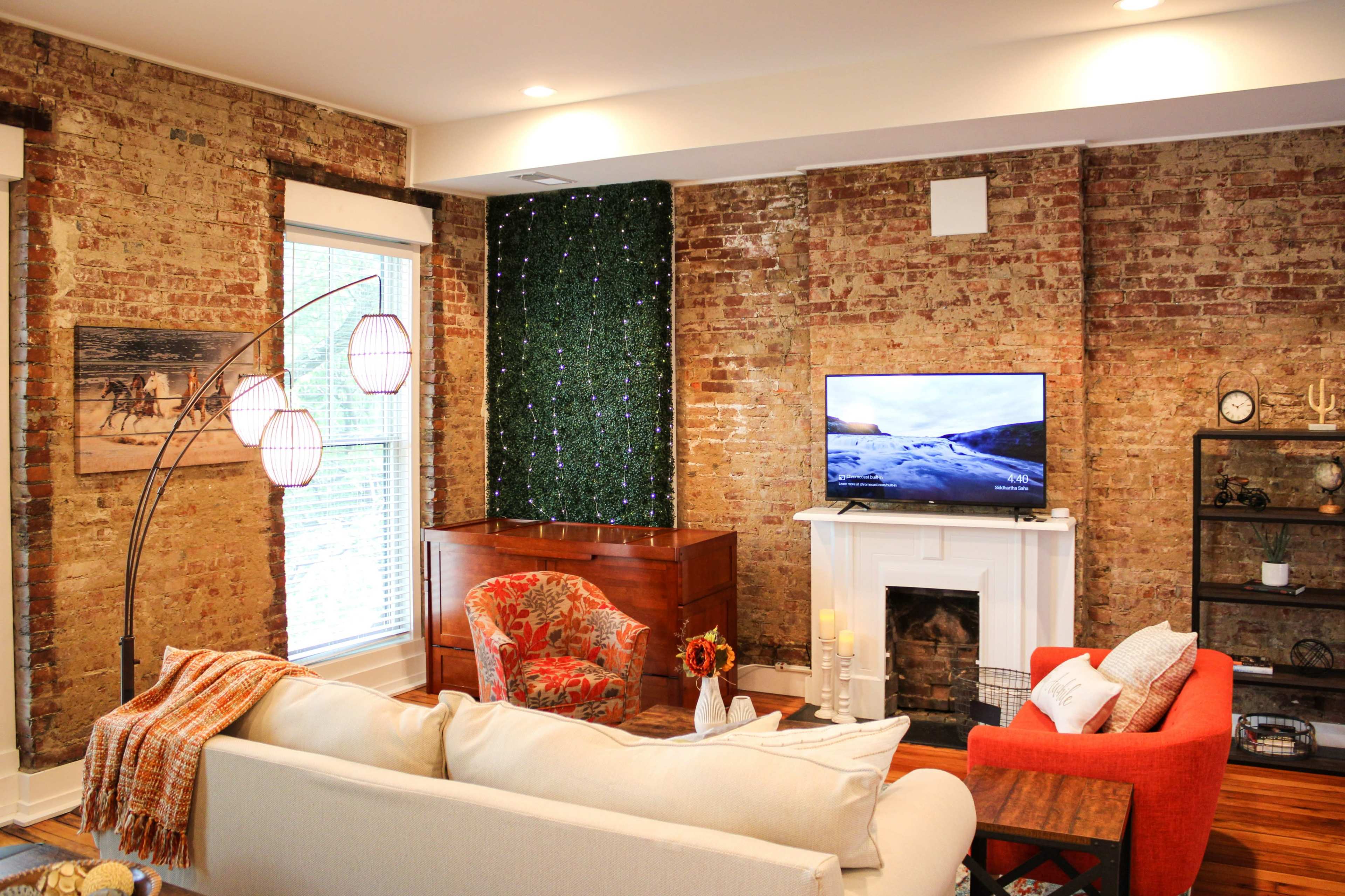A cozy living room features exposed brick walls, a television on a stand, and a sectional sofa opposite a wooden chest and a red armchair.